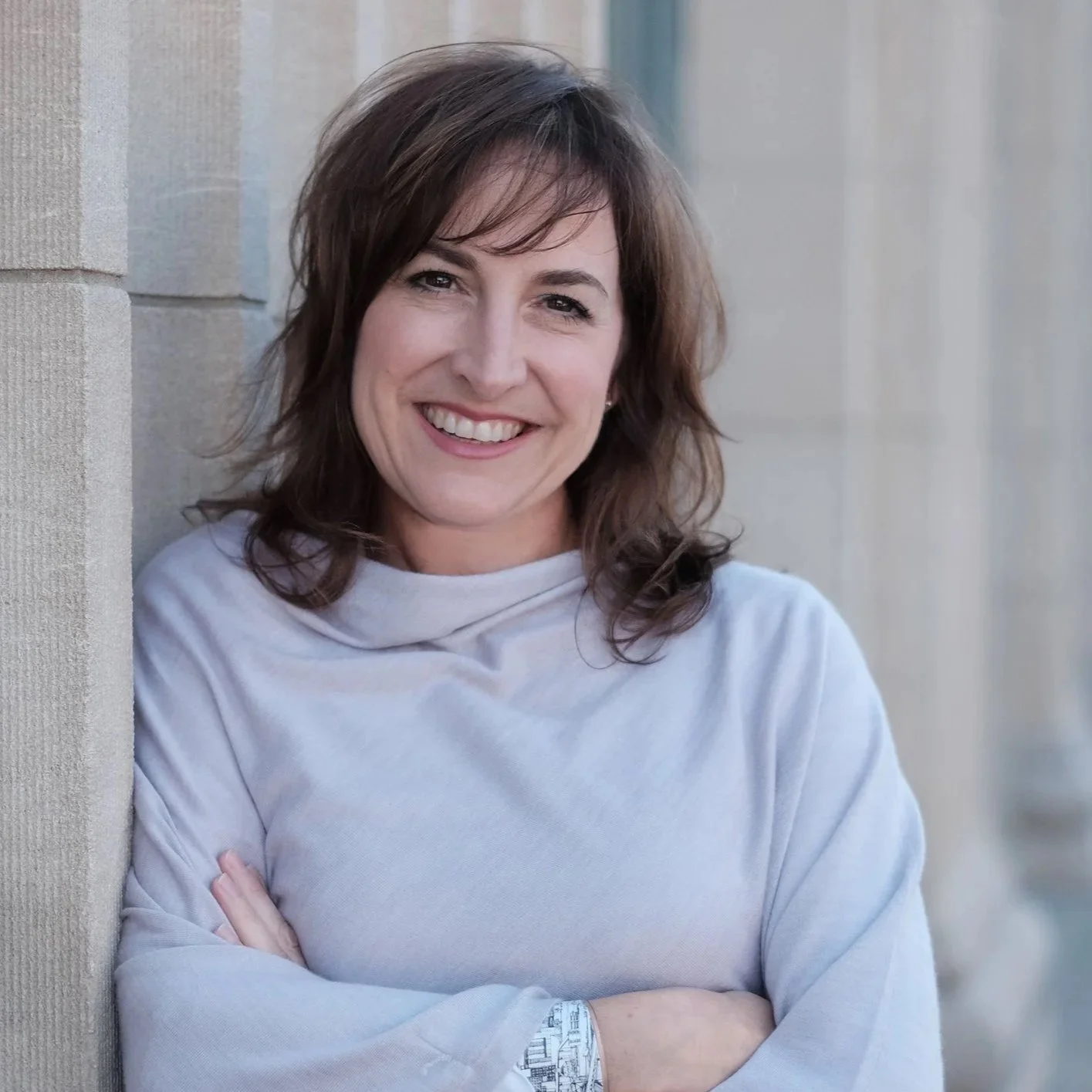 A woman with brown hair and a light-colored sweater, smiling and standing with arms crossed against a beige stone wall.