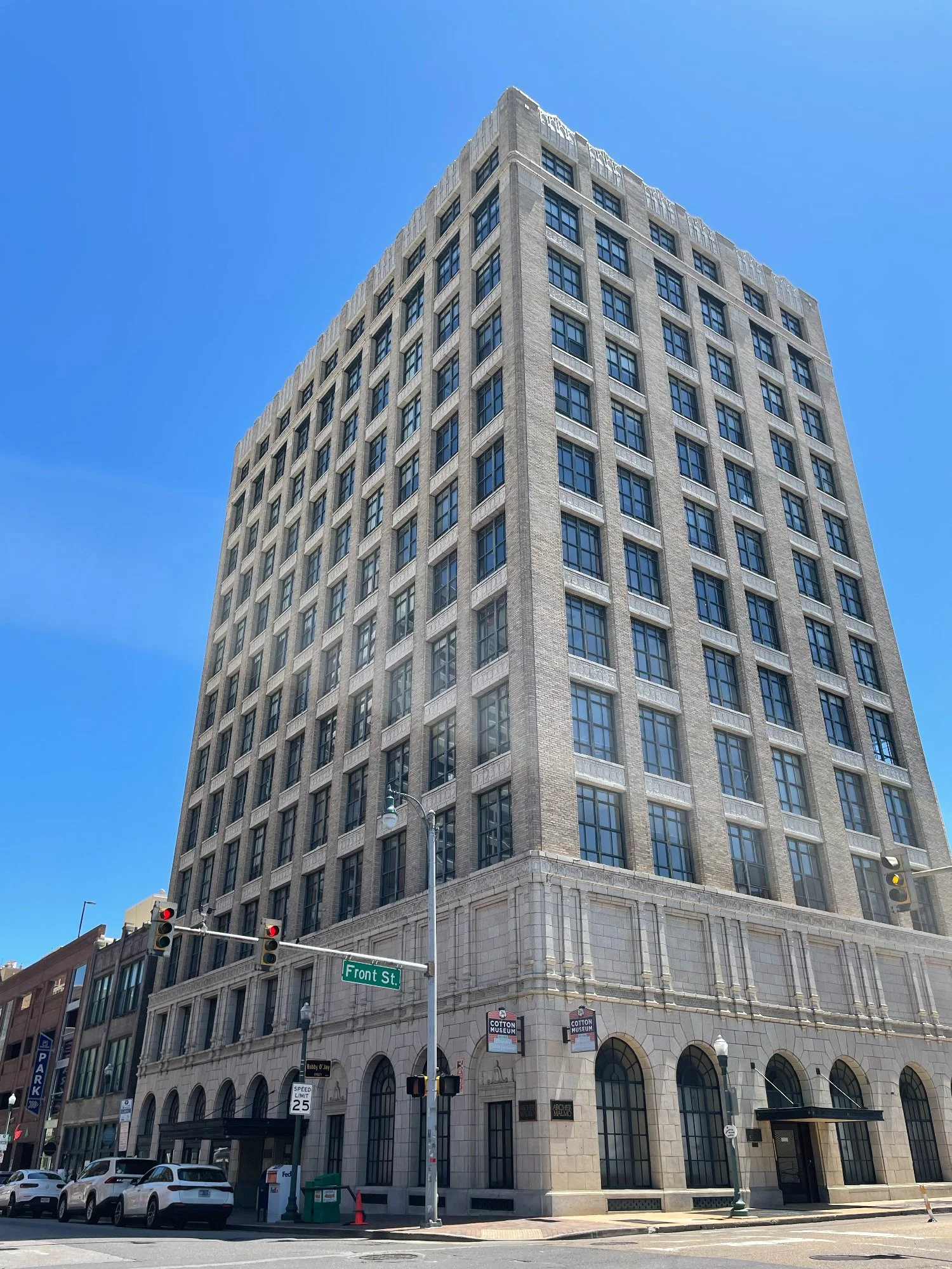 A tall, historic, multi-story building made of gray stone with numerous windows, situated on a city street corner with traffic lights, cars, and street signs. The street sign reads "Front St." and a sign indicates the Cotton Museum is nearby. The sky is clear and blue.