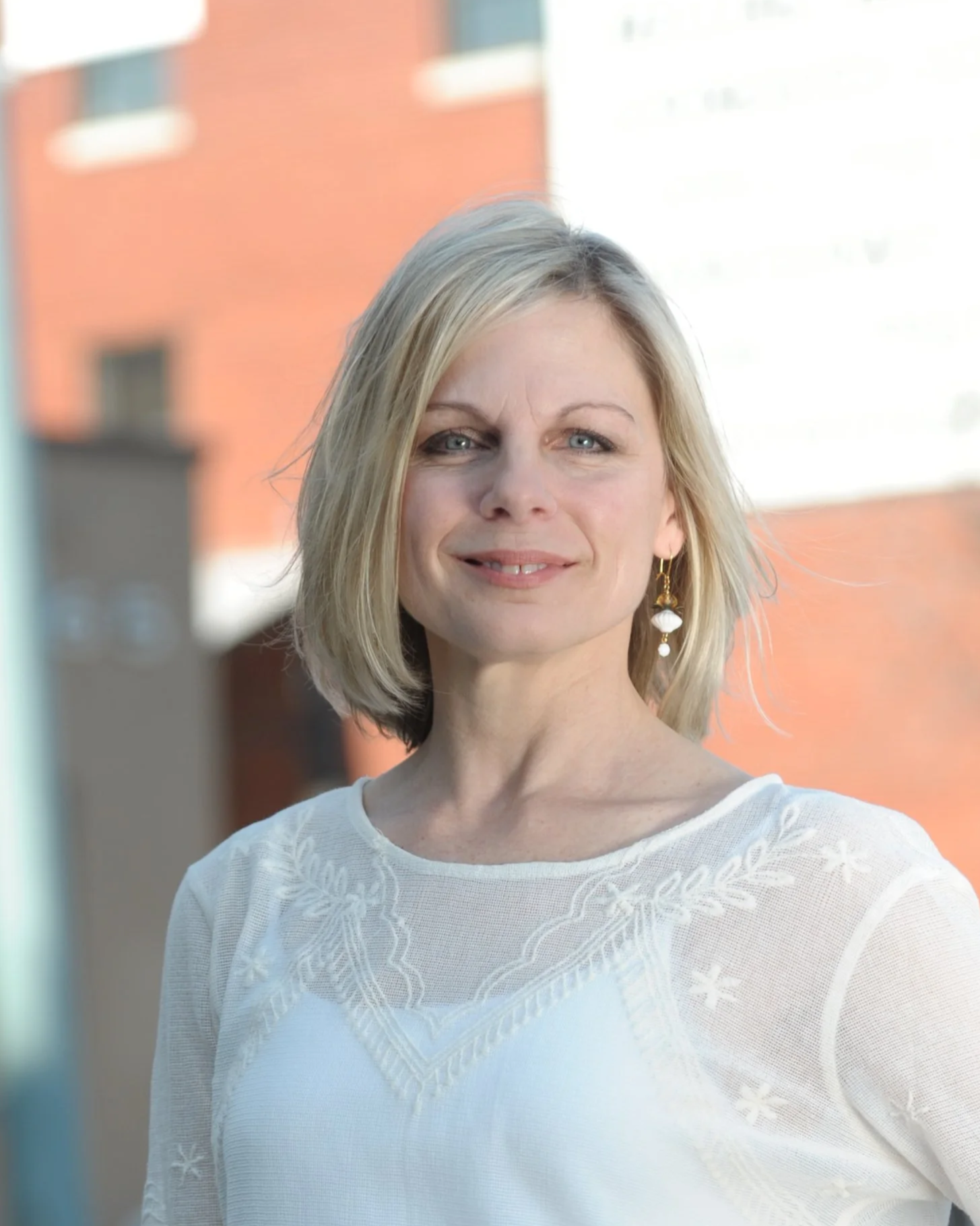 Close-up of a middle-aged woman with blonde hair, blue eyes, and earrings, standing outdoors in sunlight, with a blurred brick building in the background.