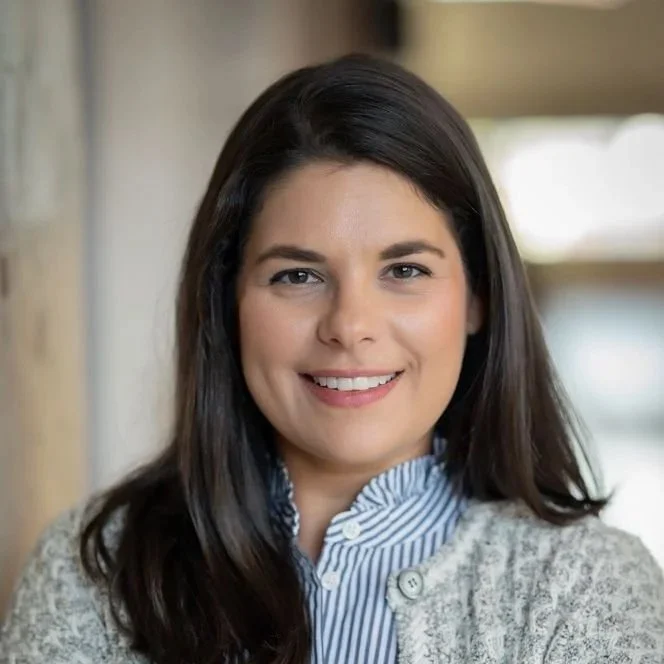 A woman with long dark hair smiling indoors, wearing a blue striped collared shirt and a gray cardigan.