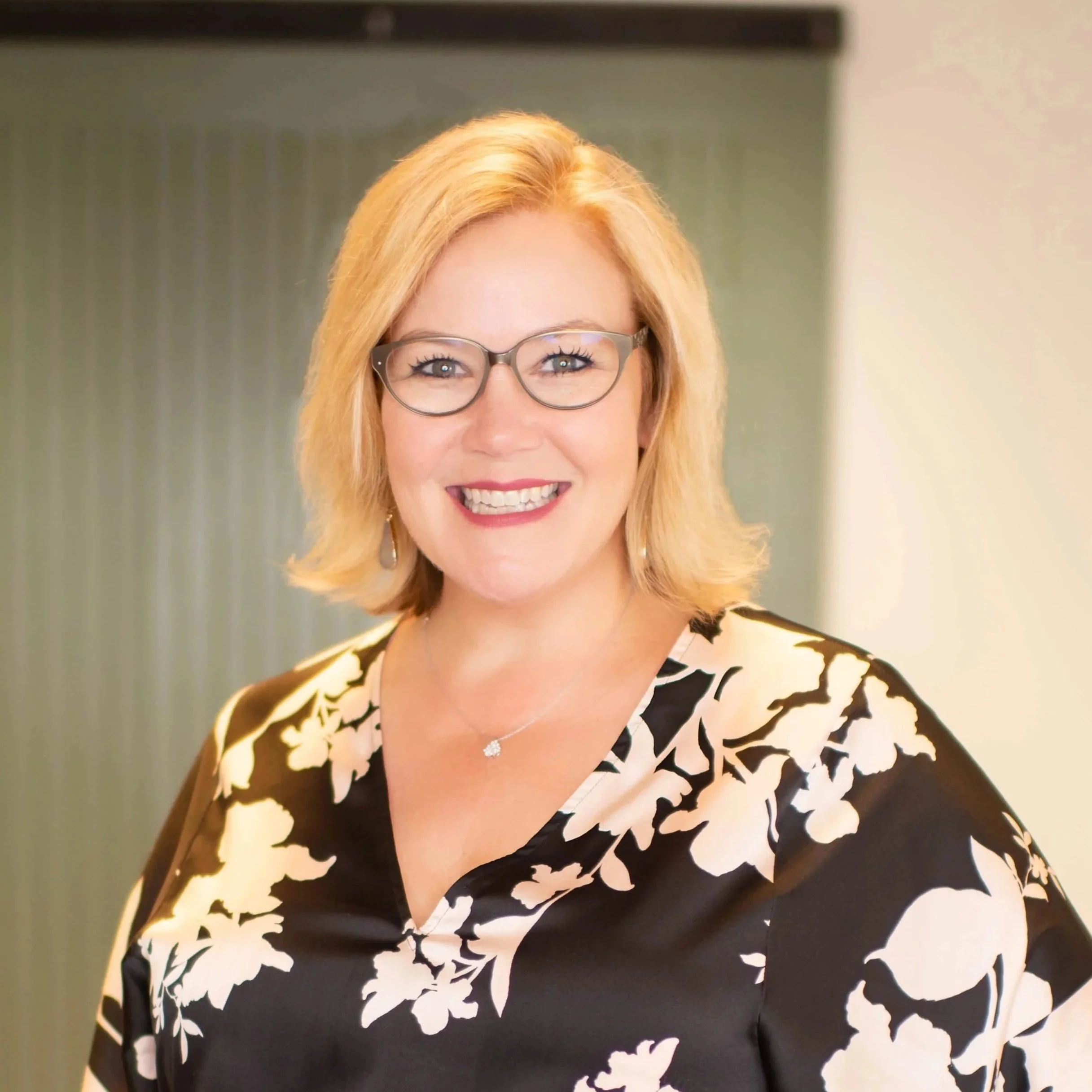 A woman with blonde hair, glasses, and a bright smile wearing a black and white floral-patterned dress.