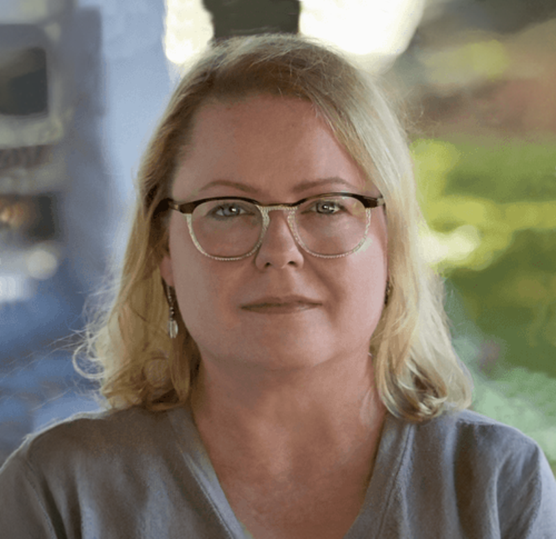 A middle-aged woman with blonde hair wearing glasses and a gray top, sitting outdoors with blurred greenery in the background.