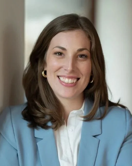 A woman with shoulder-length brown hair, wearing a light blue blazer, white shirt, gold hoop earrings, smiling, in an indoor setting with a blurred background.