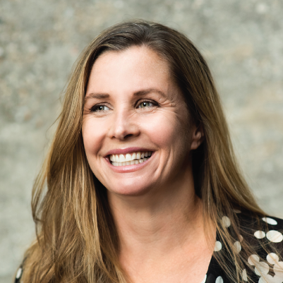 A woman with long brown hair and light skin smiling, wearing a dark polka dot top, against a neutral background.