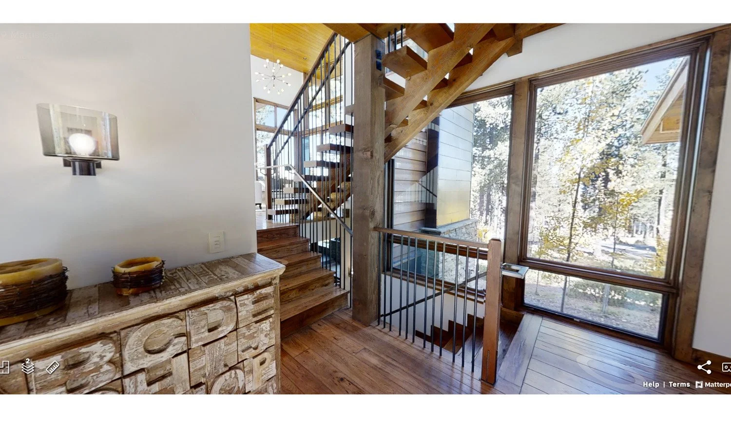 Wooden staircase with metal railings and large windows showing outdoor scenery, interior decorated with modern fixtures and a rustic-style cabinet.