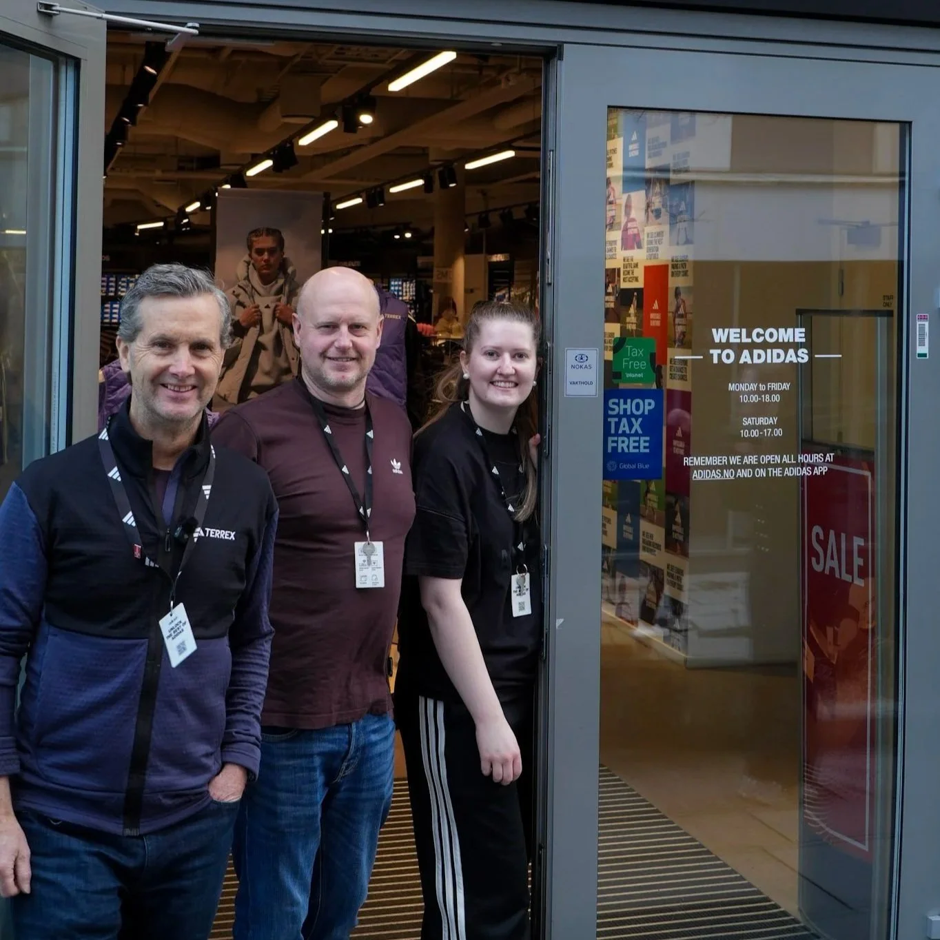 Tre ansatte står utenfor en Adidas-butikk, smilende og ser mot kamera. To av dem har på seg Adidas klær.