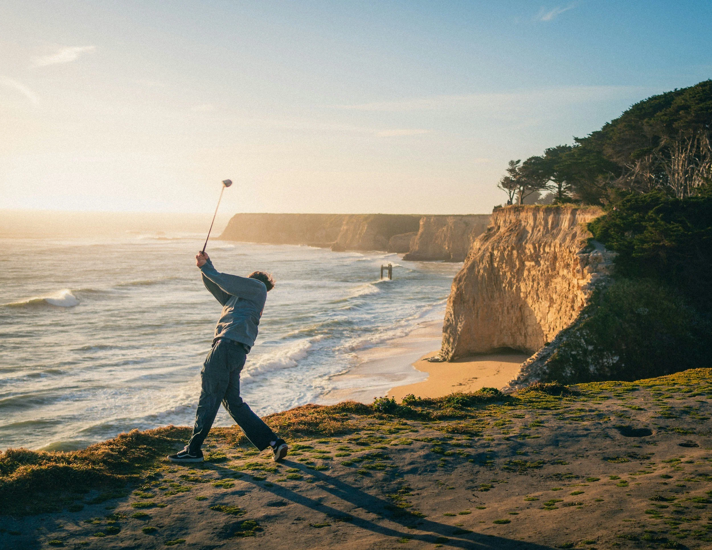A person finishing a golf swing on a coastal cliffs overlooking the ocean at sunset.