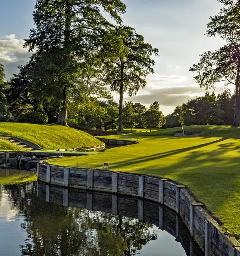 A golf course with well-maintained green grass, a small water hazard with reflections, surrounded by trees and a partly cloudy sky.