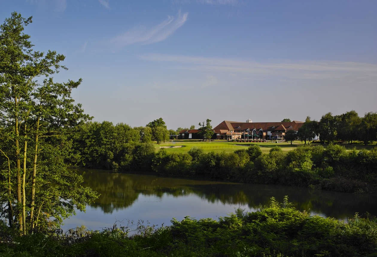 A scenic view of a golf course with a large building in the background, surrounded by trees and a river in the foreground, under a clear blue sky.