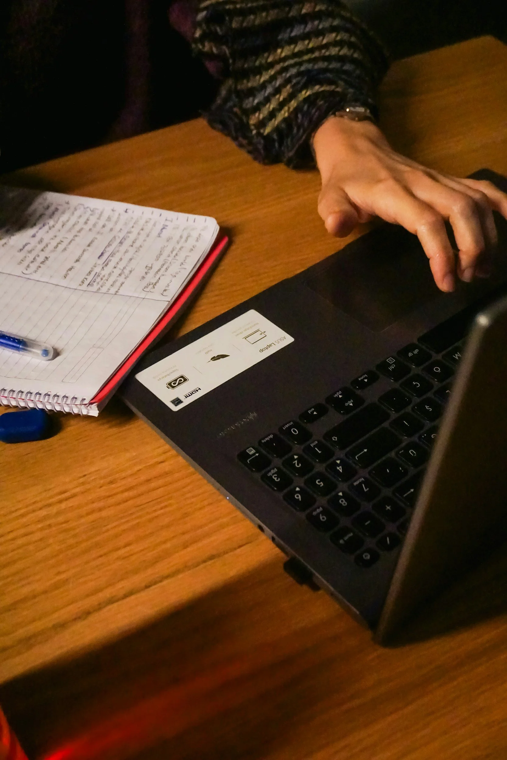 Close-up of a person's hand resting on a closed laptop on a wooden table, with an open notebook and a pen nearby.