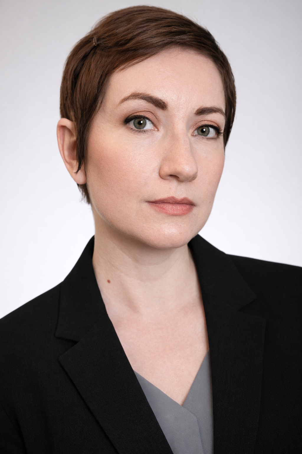 A professional woman with short red hair and light makeup, wearing a black blazer over a gray top, against a plain white background.