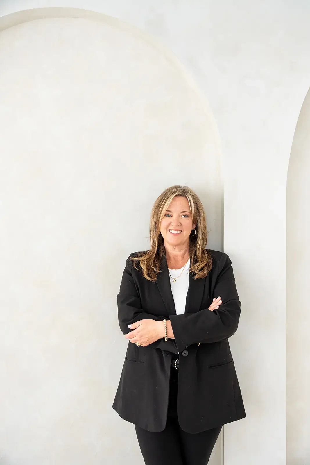 A woman with long hair wearing a black blazer and white shirt stands with arms crossed, smiling, in front of a white arched wall.