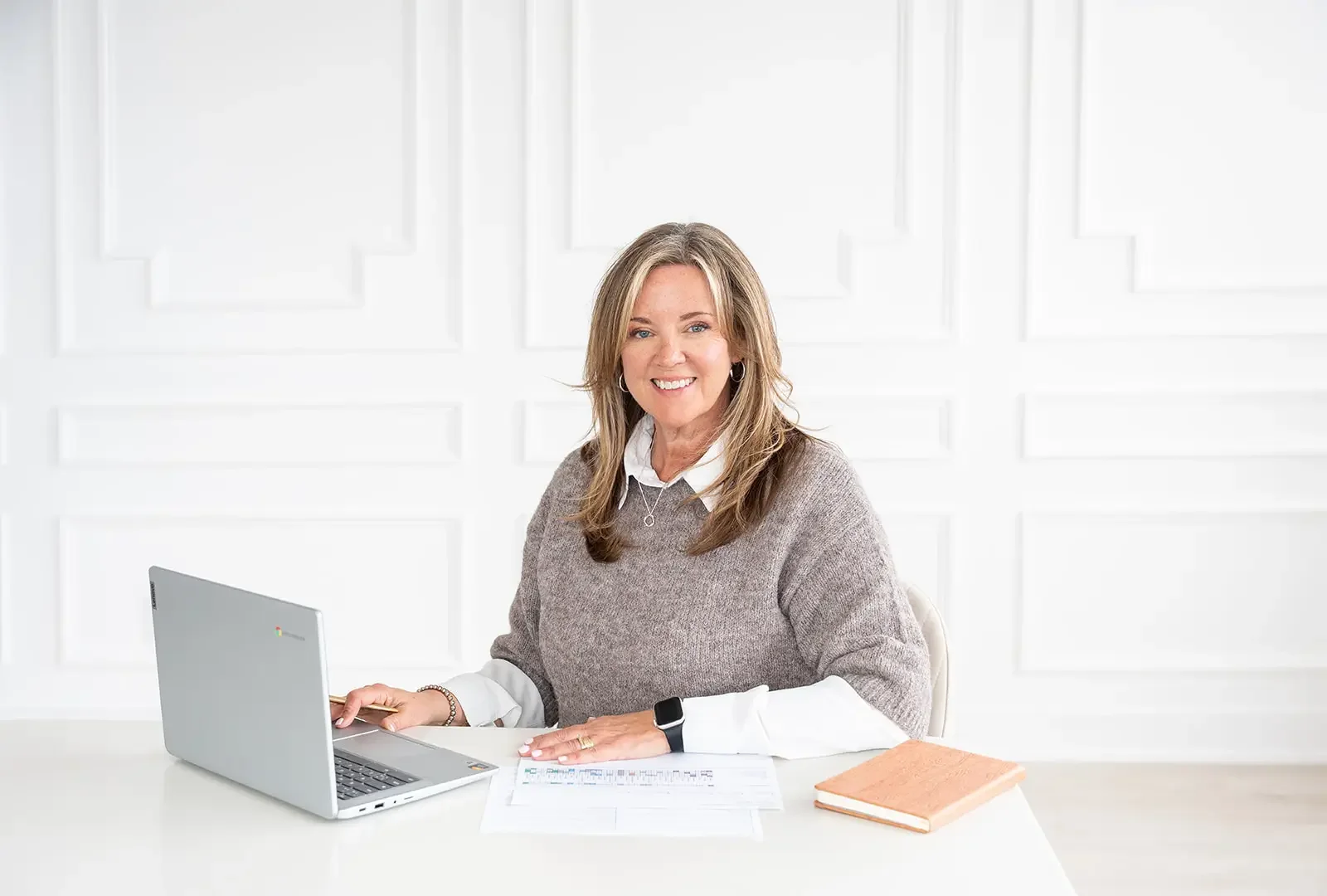 Woman sitting at white desk with a silver laptop, documents, and a closed notebook, smiling toward the camera in a bright, white room