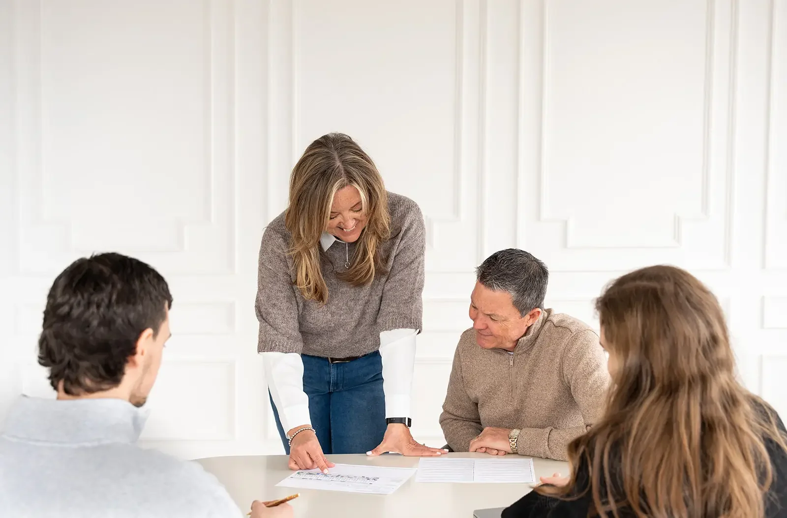 A woman standing and smiling at a group of four seated people, looking at a document on a table.