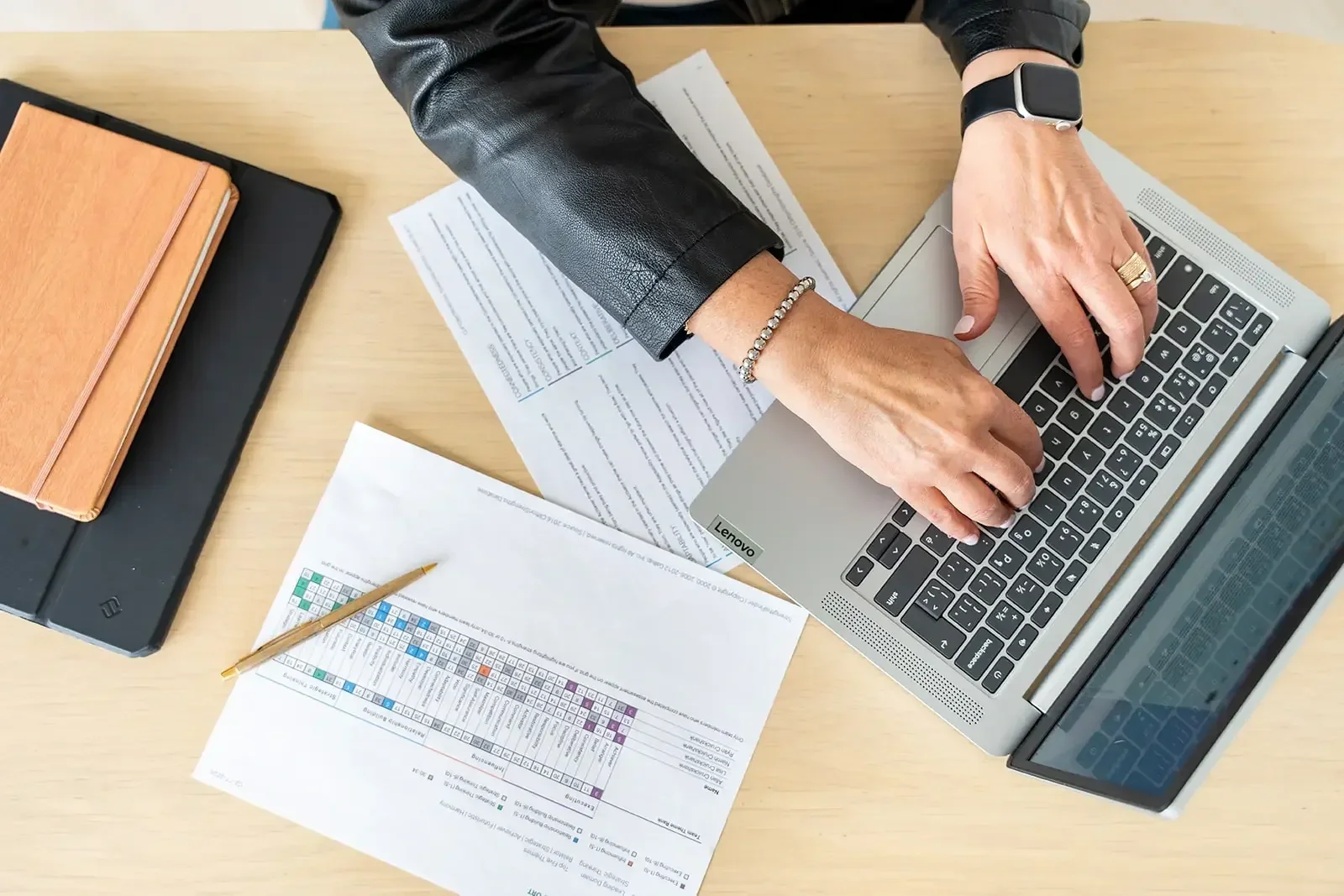 Person working on a laptop at a desk with papers, notebooks, and pens.