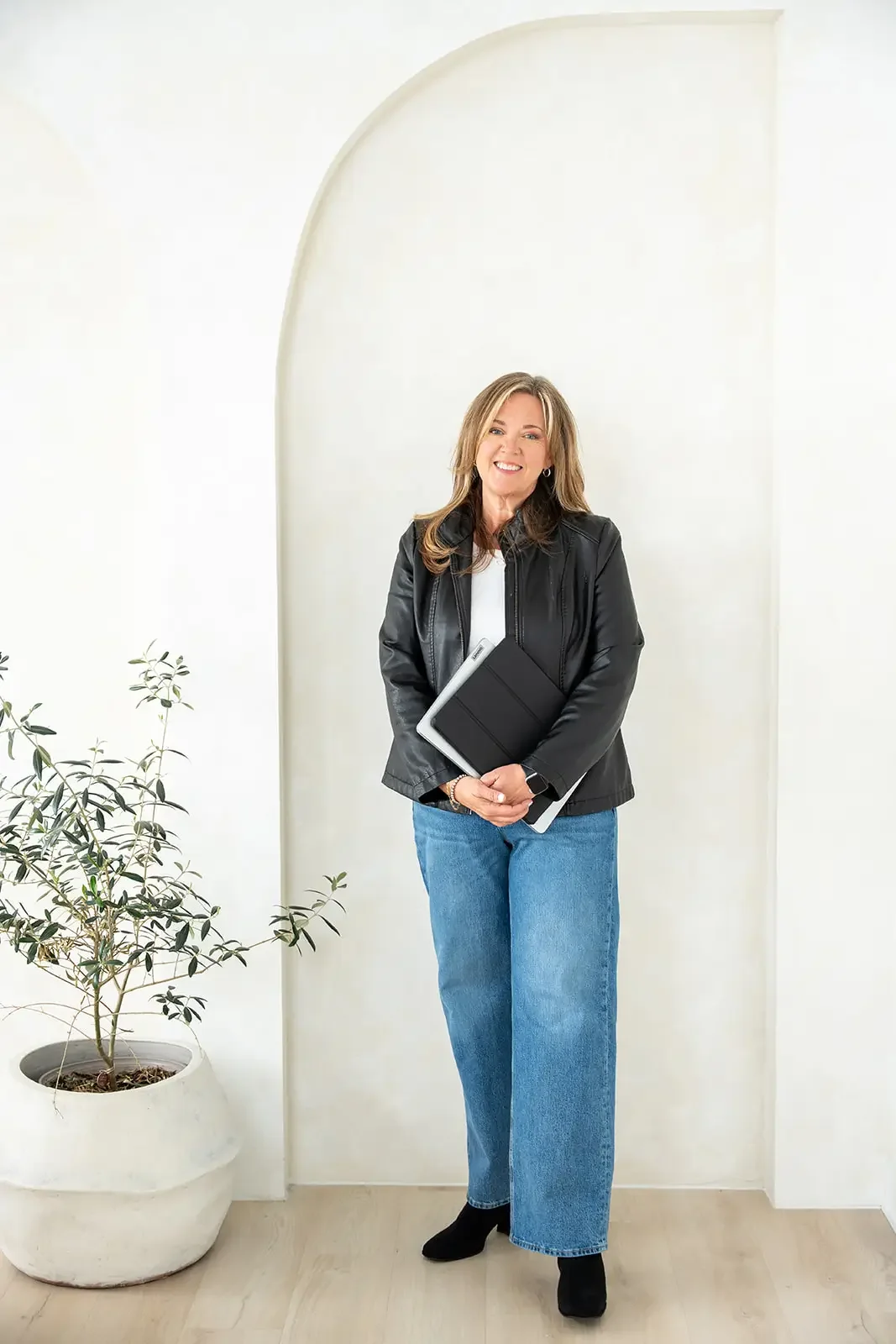 A smiling woman with light brown hair wearing a black leather jacket, white top, and blue jeans, holding a tablet, standing indoors near a potted plant with a white wall and an arch in the background.