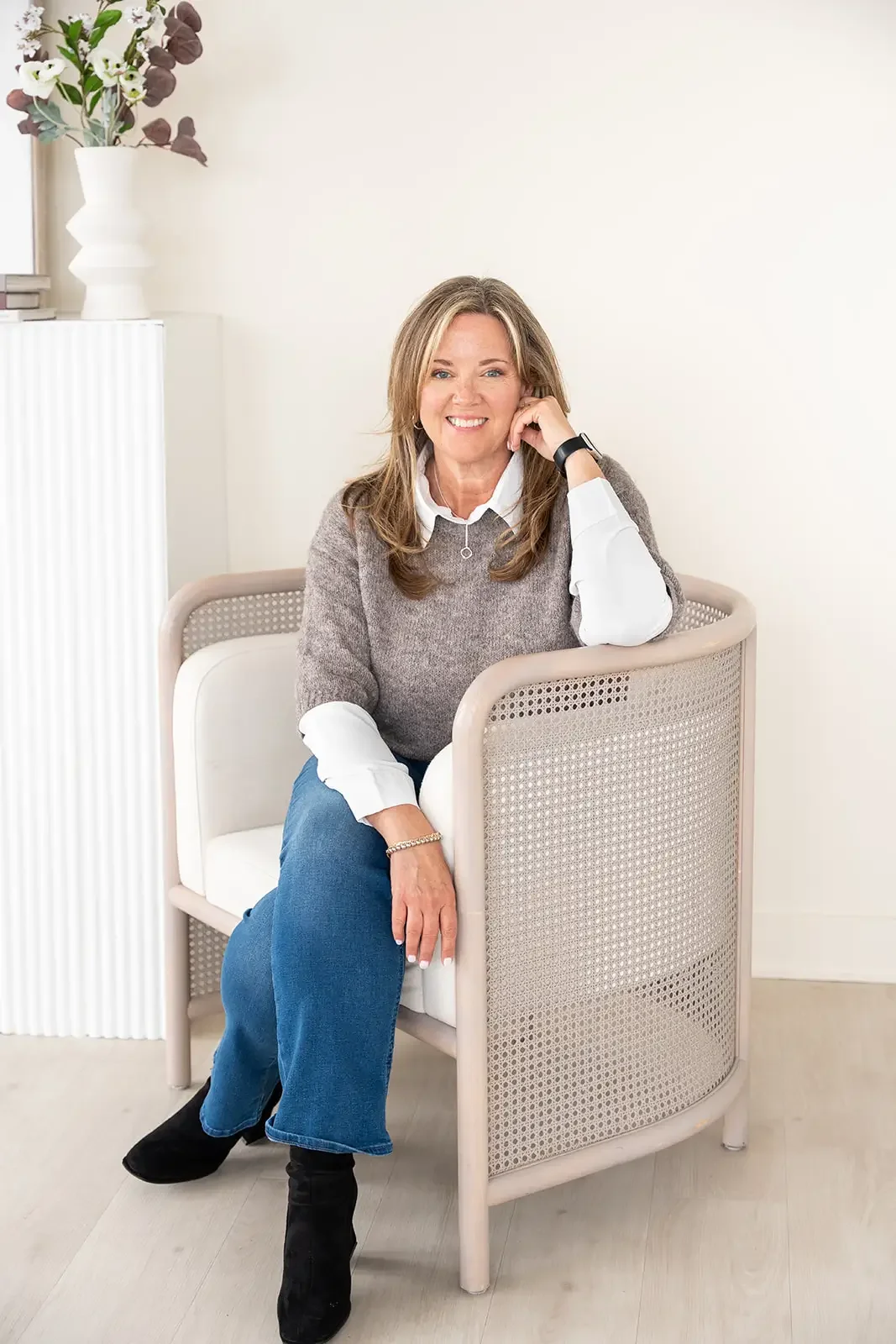 Smiling woman sitting in a beige chair with rattan sides, wearing a white shirt under a gray sweater, blue jeans, and black boots, in a bright room with a white wall, a white radiator, and a vase with flowers on a white cabinet in the background.