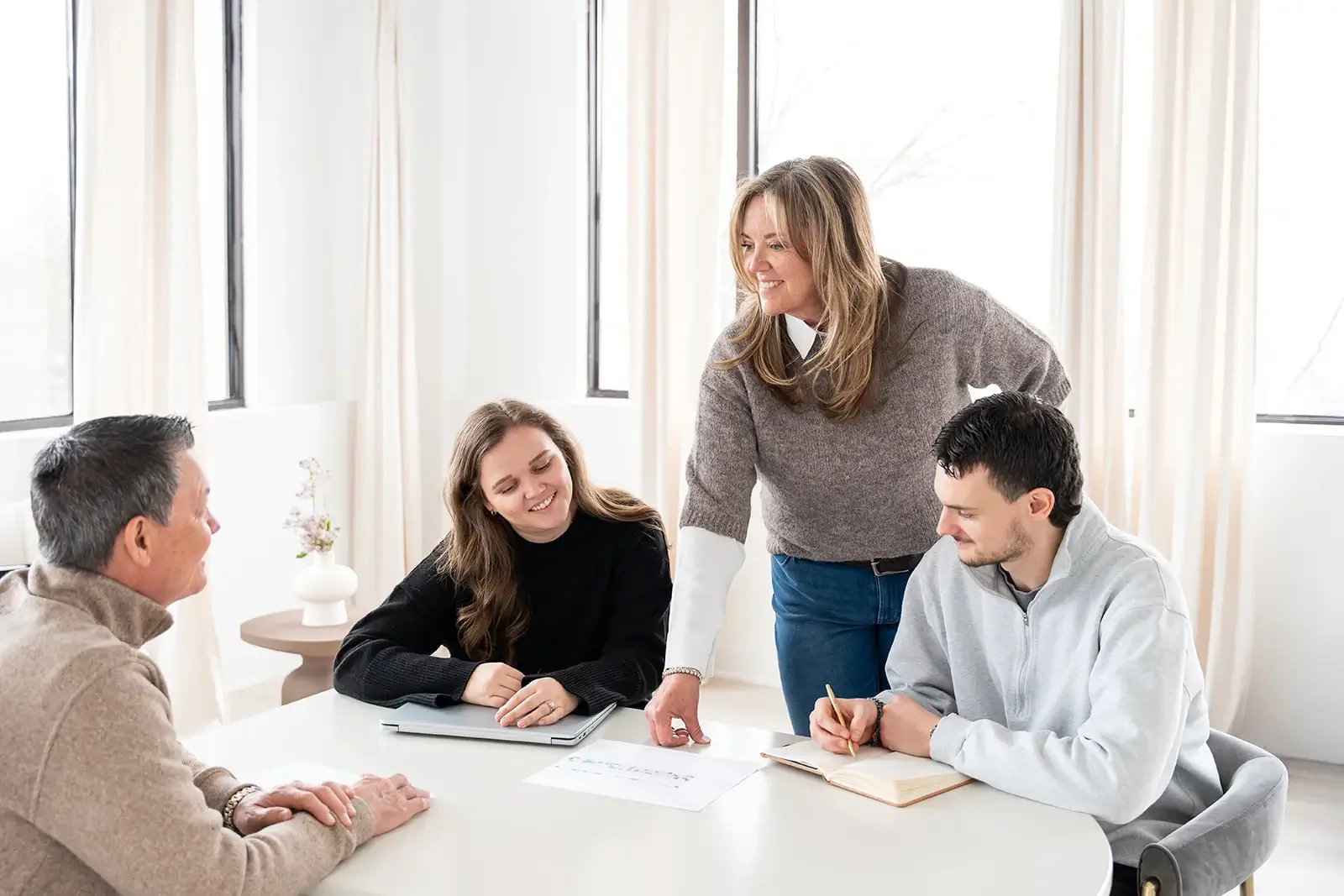 Four people engaged in a discussion at a meeting, with one woman standing and speaking, while three seated individuals look on with smiles.