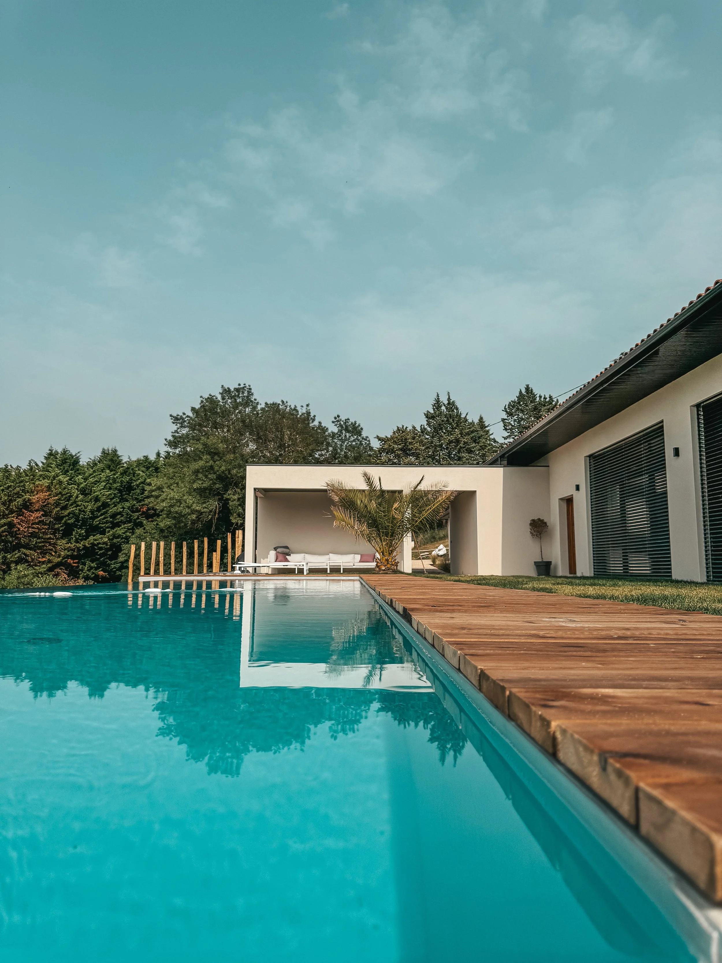 Une piscine de bord en bois avec une maison moderne en arrière-plan, entourée d'arbres et d'un ciel clair avec quelques nuages.
