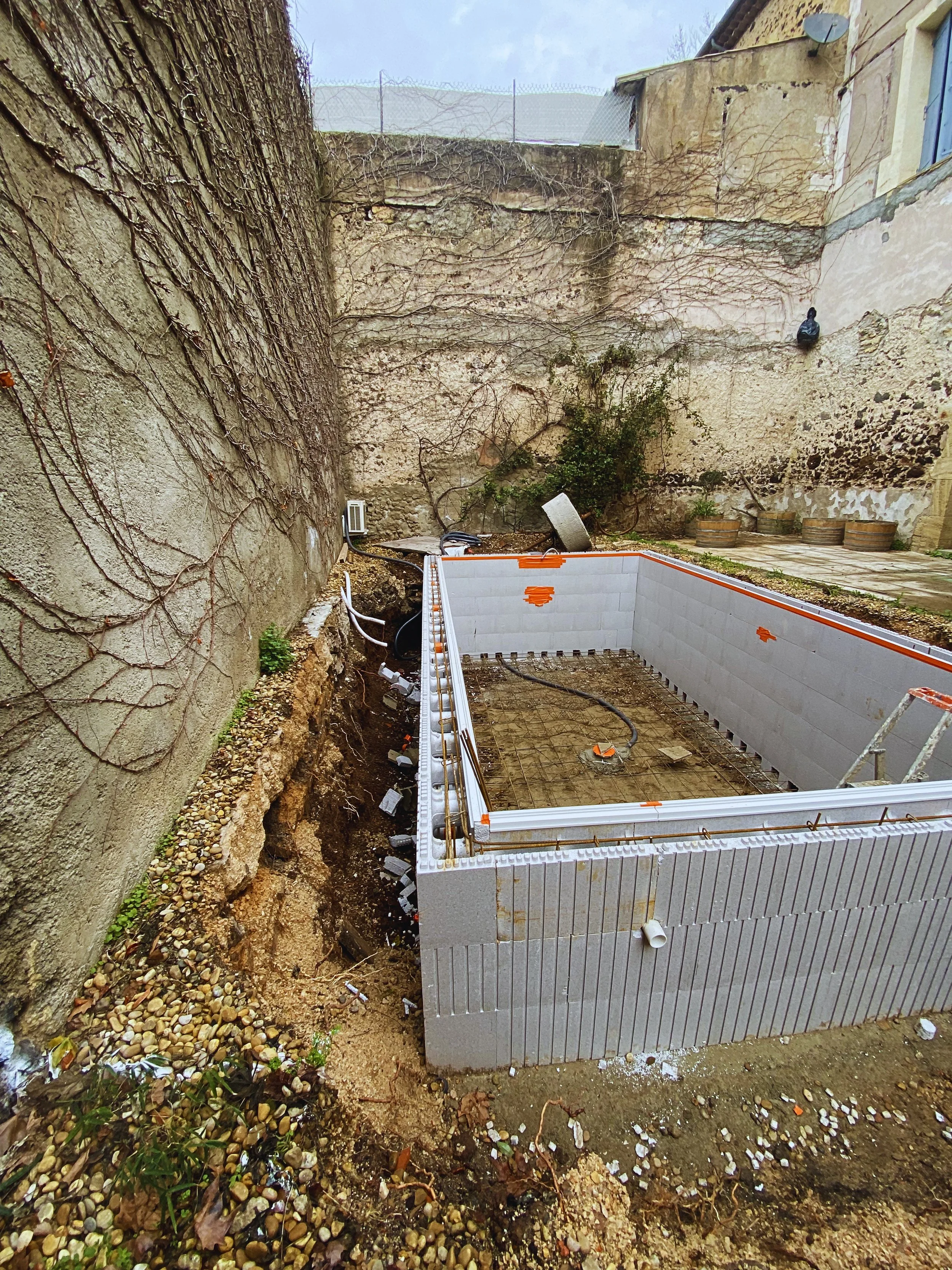 Construction d'une piscine en béton dans une cour entourée de vieux murs en pierre, avec des outils et matériaux de construction autour.