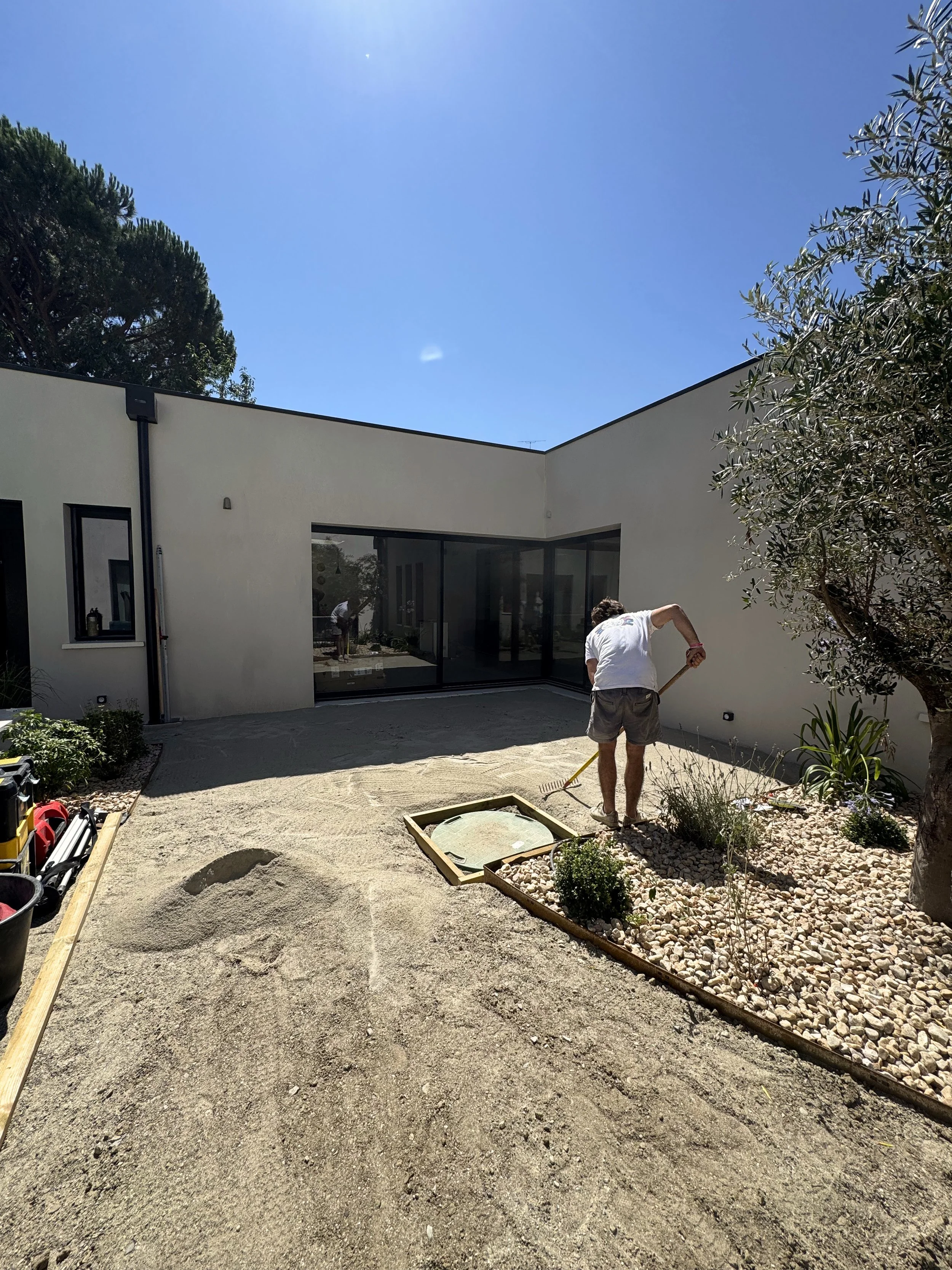 Un homme travaille dans une cour extérieure de maison moderne, avec un jardin planté de petites plantes et un massif de galets. La maison a de grandes fenêtres et des murs blancs, sous un ciel clair et ensoleillé.