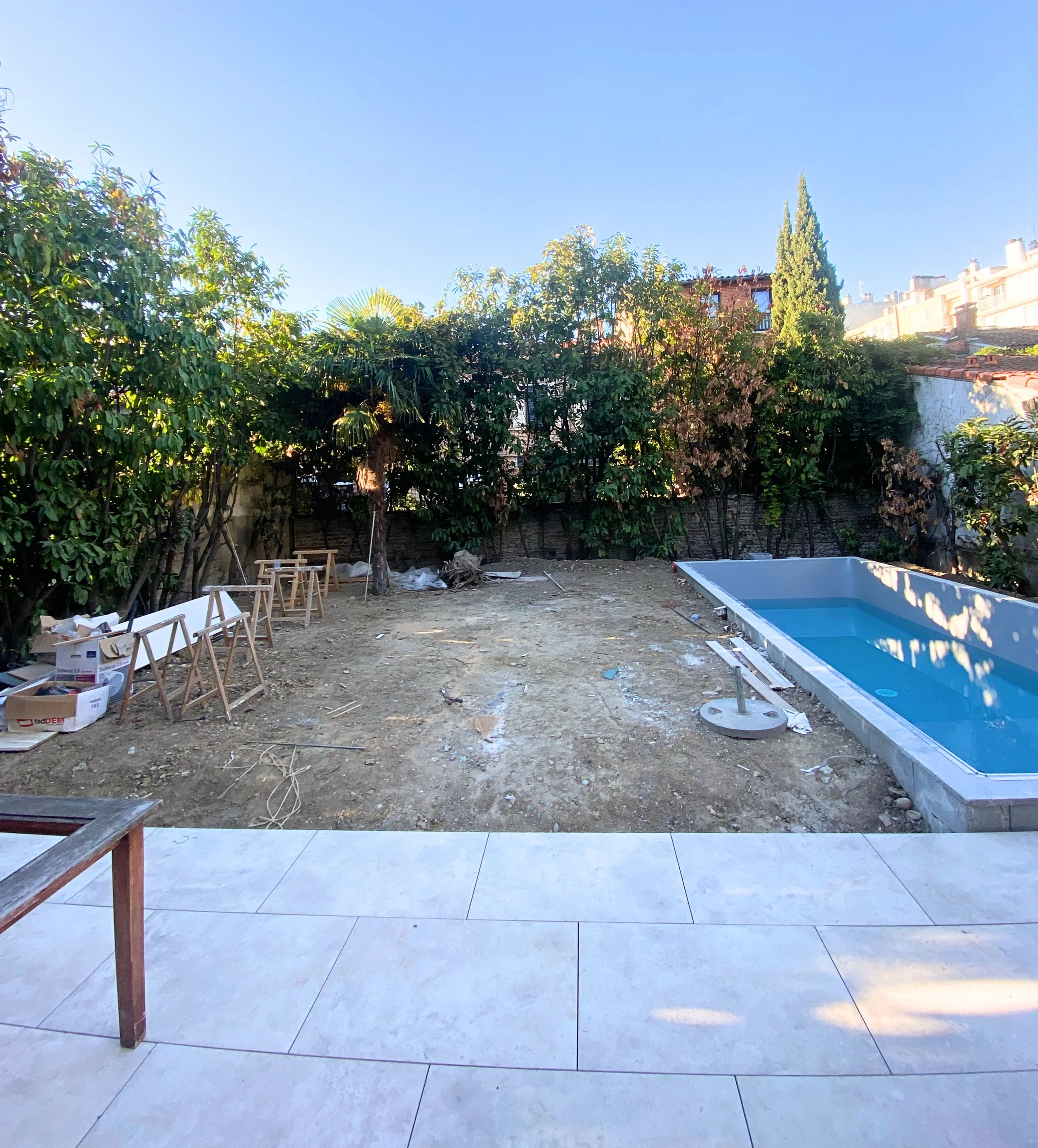 Espace extérieur avec sol en carreaux, piscine vide en construction, zone avec terre et déchets, végétation dense en arrière-plan, et une partie de terrasse en premier plan.