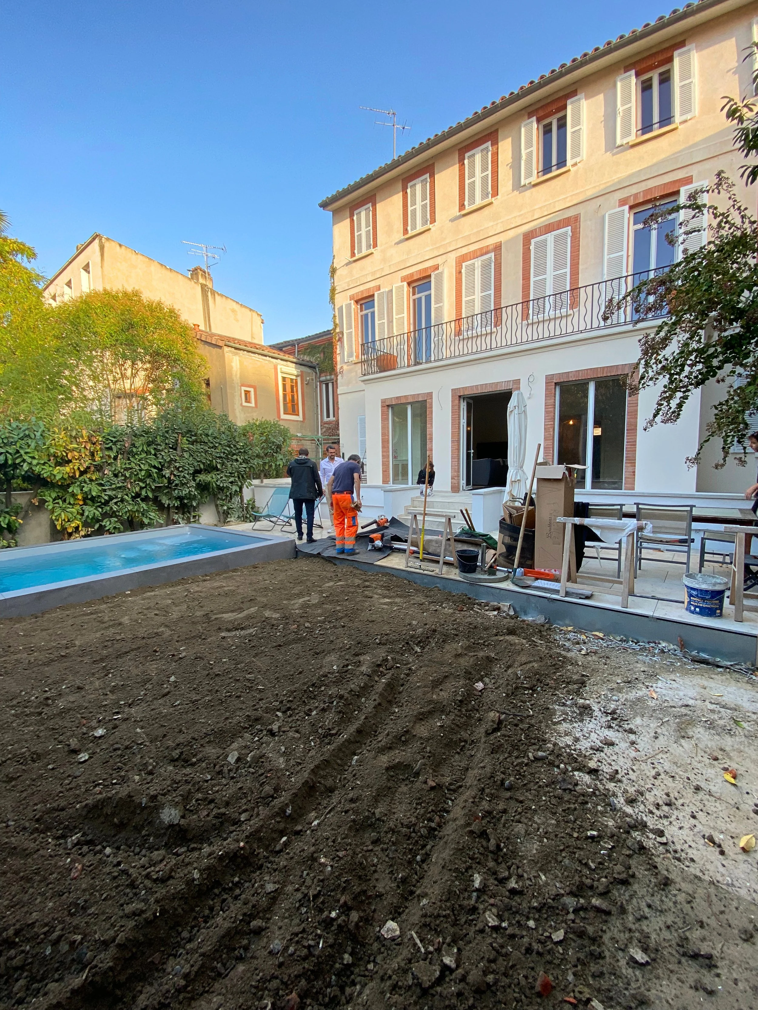 Une cour avec un bâtiment résidentiel à plusieurs étages en arrière-plan, des ouvriers effectuant des travaux de construction ou de rénovation, des outils et des meubles empilés, un petit bassin ou piscine, et un terrain en terre non encore travaillé.
