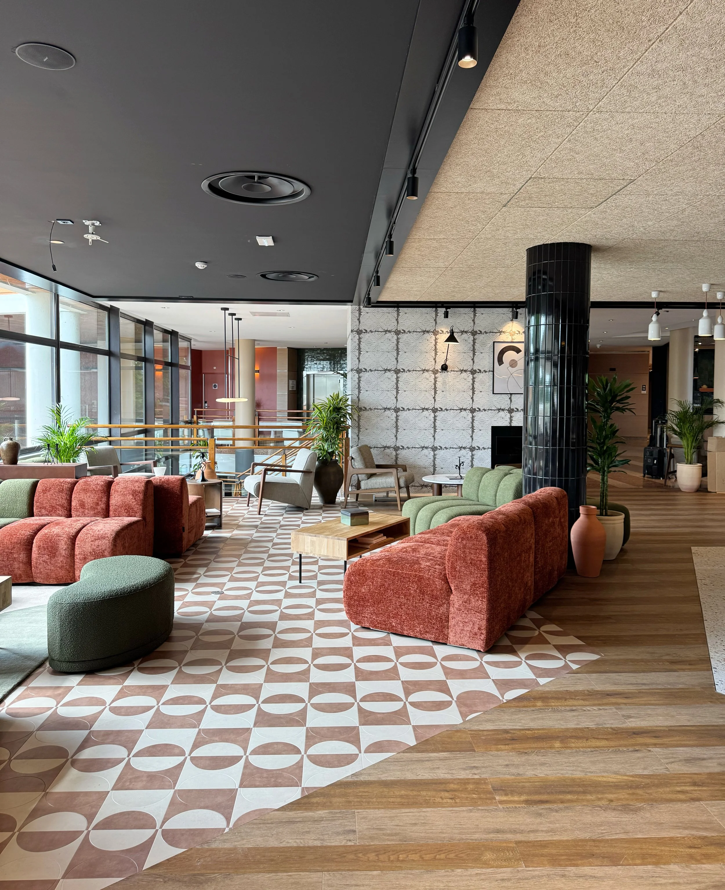Intérieur moderne d'un hall d'hôtel ou de lounge avec des sièges en velours rouge et vert, des plantes, un sol en carreaux de céramique avec un motif géométrique, des murs en mosaïque et en briques visibles, ainsi que des éléments de décoration comme des cadres, des lampes pendantes et des vases en terre cuite.