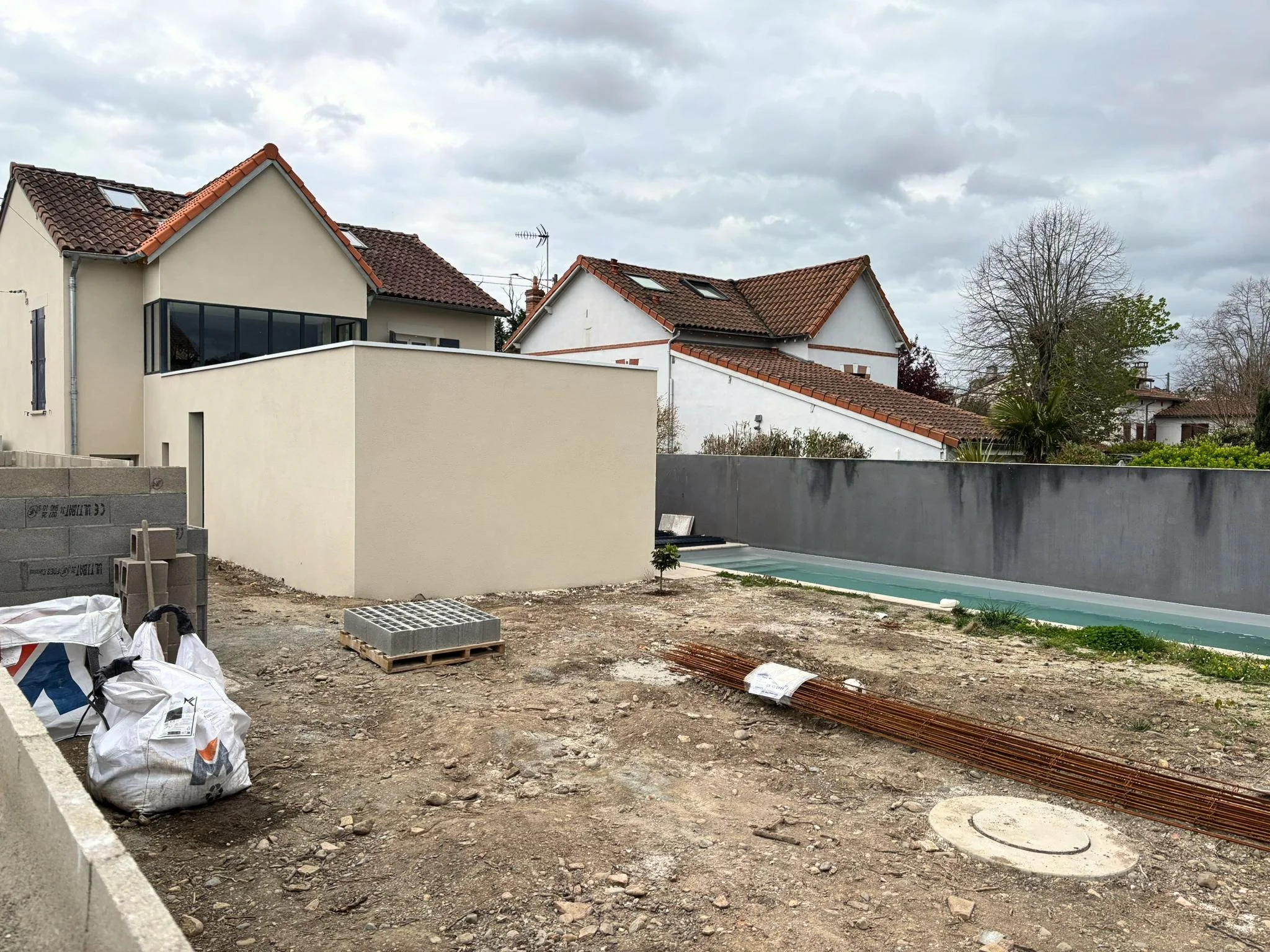 Construction de maison avec des matériaux comme des parpaings, des tiges de fer, et des sacs de ciment, avec un terrain en terre et des maisons avec des toits en tuiles dans le fond.
