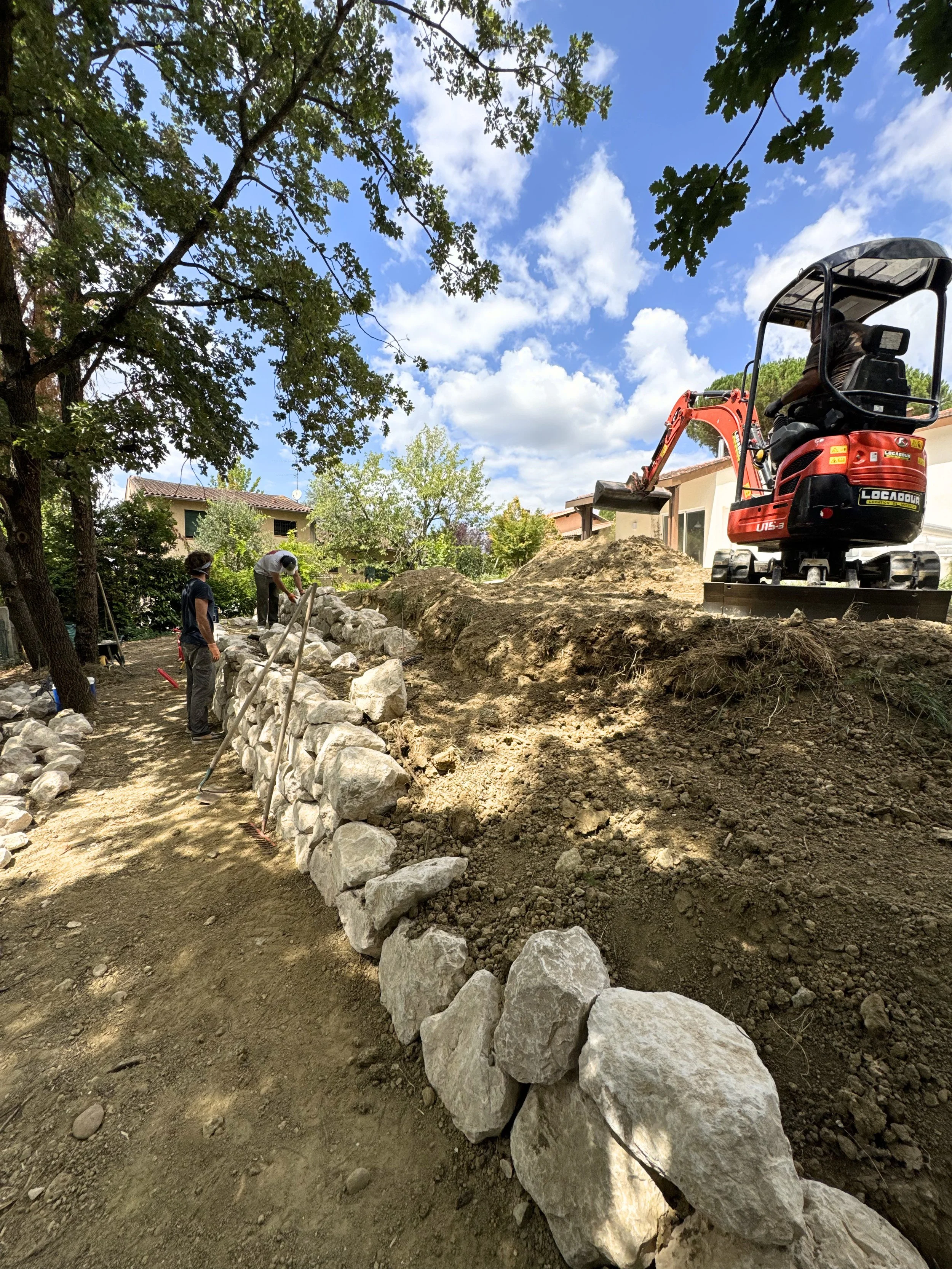 Travaux de construction avec un mur de pierres, une petite pelleteuse rouge, et des ouvriers travaillant en plein air sous un ciel bleu avec des nuages, dans un environnement résidentiel avec des arbres et des maisons.
