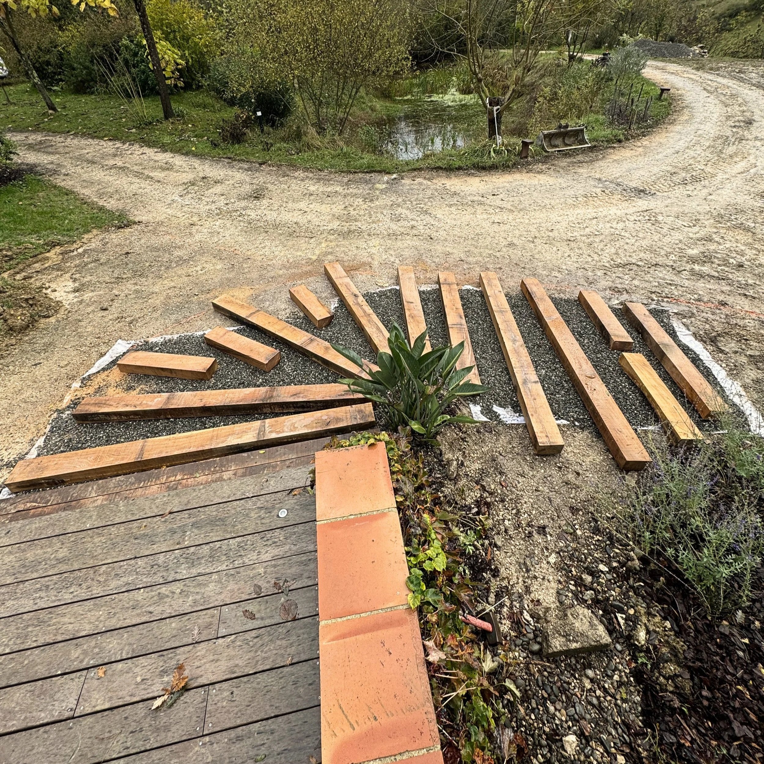 Un chemin de terre en courbe dans un parc avec des arbres, une petite mare et une brouette abandonnée à côté de l'eau, vue partielle de la scène. En premier plan, un assemblage de planches en bois déposé au sol.