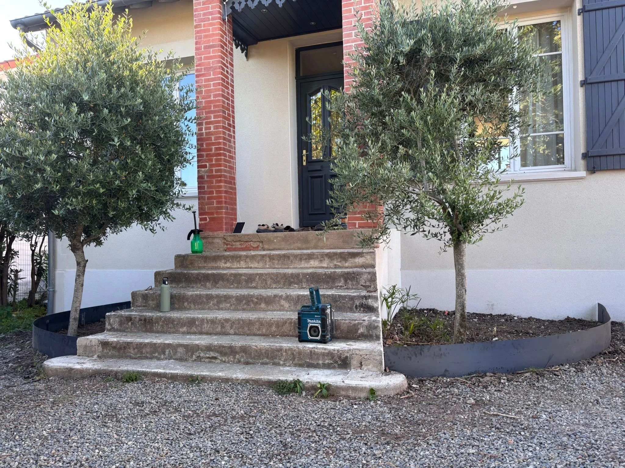 Une maison avec une entrée principale, deux arbres de chaque côté, des marches en pierre menant à la porte, et des outils de travail au sol