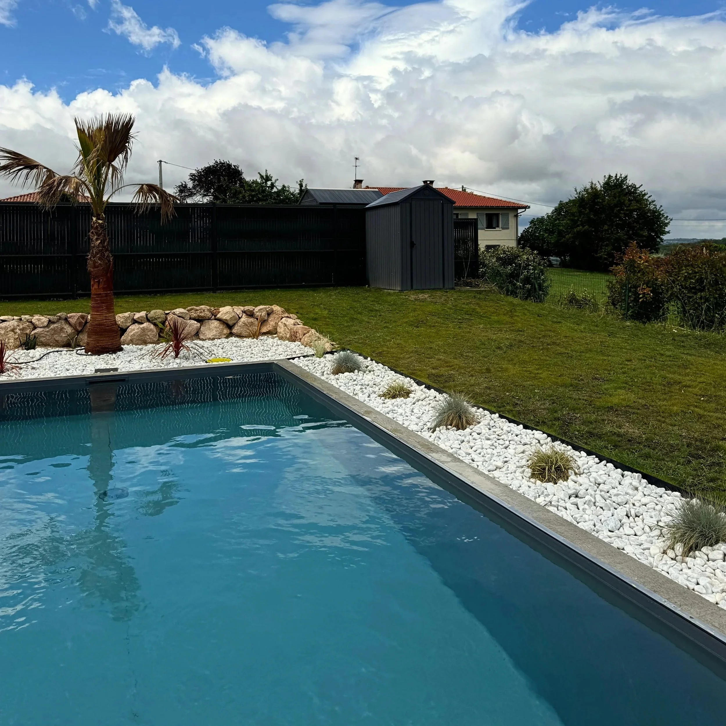 Une vue d'une piscine avec un jardin entouré de pierres blanches et une clôture en métal noire derrière, un palmier en premier plan et des maisons avec des toits en tuiles rouges et des arbres en arrière-plan, sous un ciel partiellement couvert de nuages.