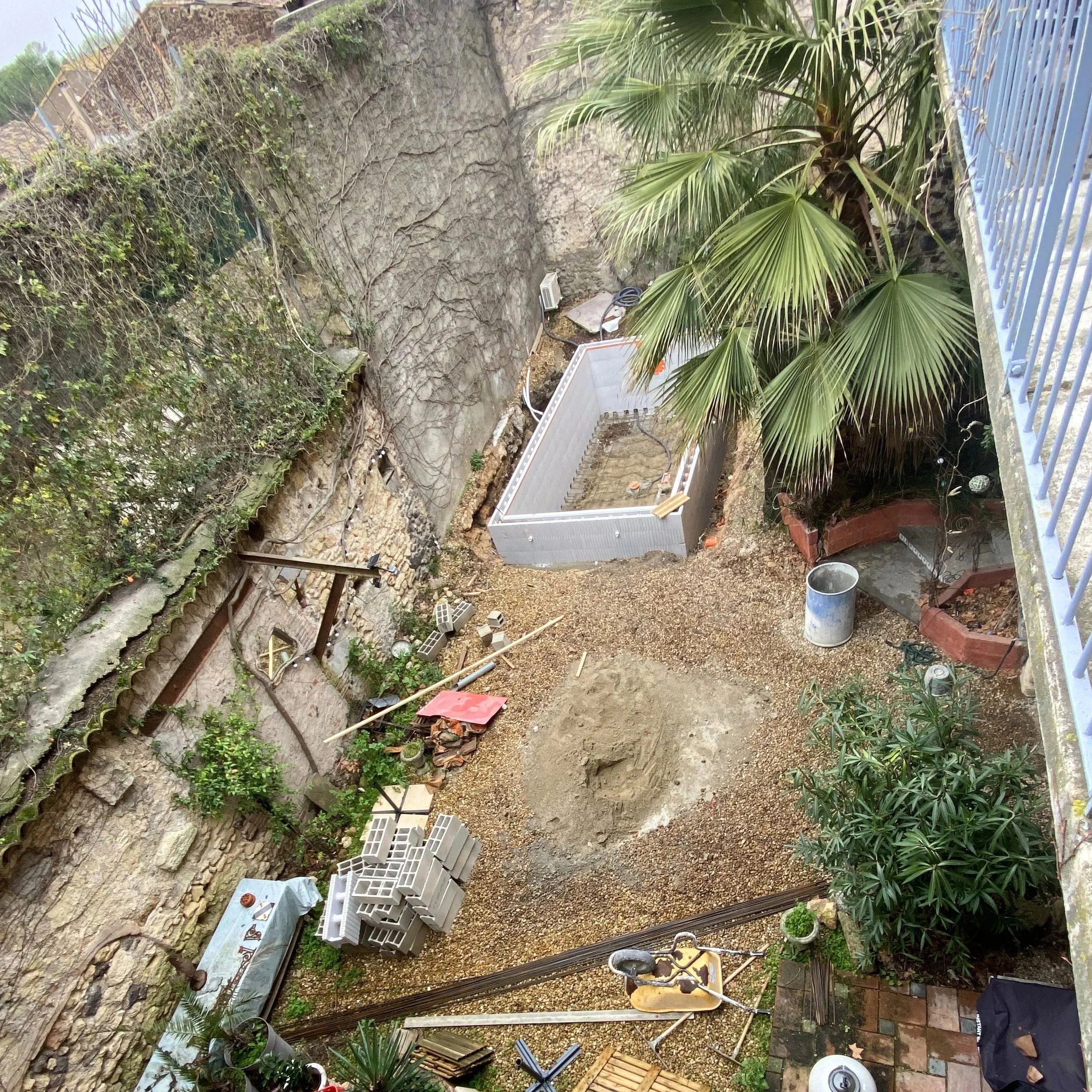 Un espace extérieur en cours de construction ou de rénovation avec des blocs de béton empilés, des plantes tropicales, un mur en pierre, du matériel de construction, et divers outils et matériaux dispersés au sol.
