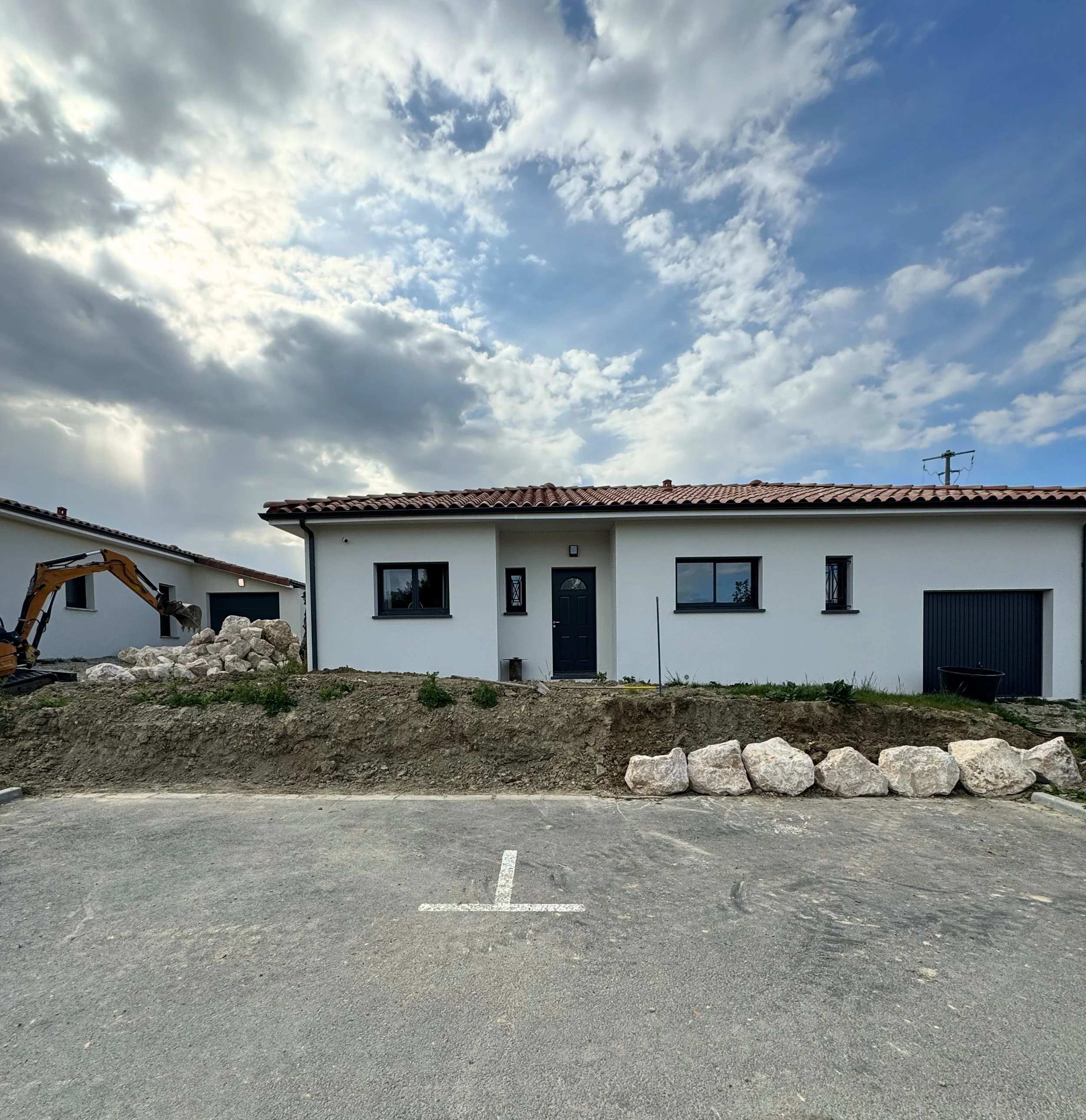 Une maison blanche avec un toit en tuiles rouges, vue depuis un parking vide avec des rochers au premier plan et un ciel nuageux.
