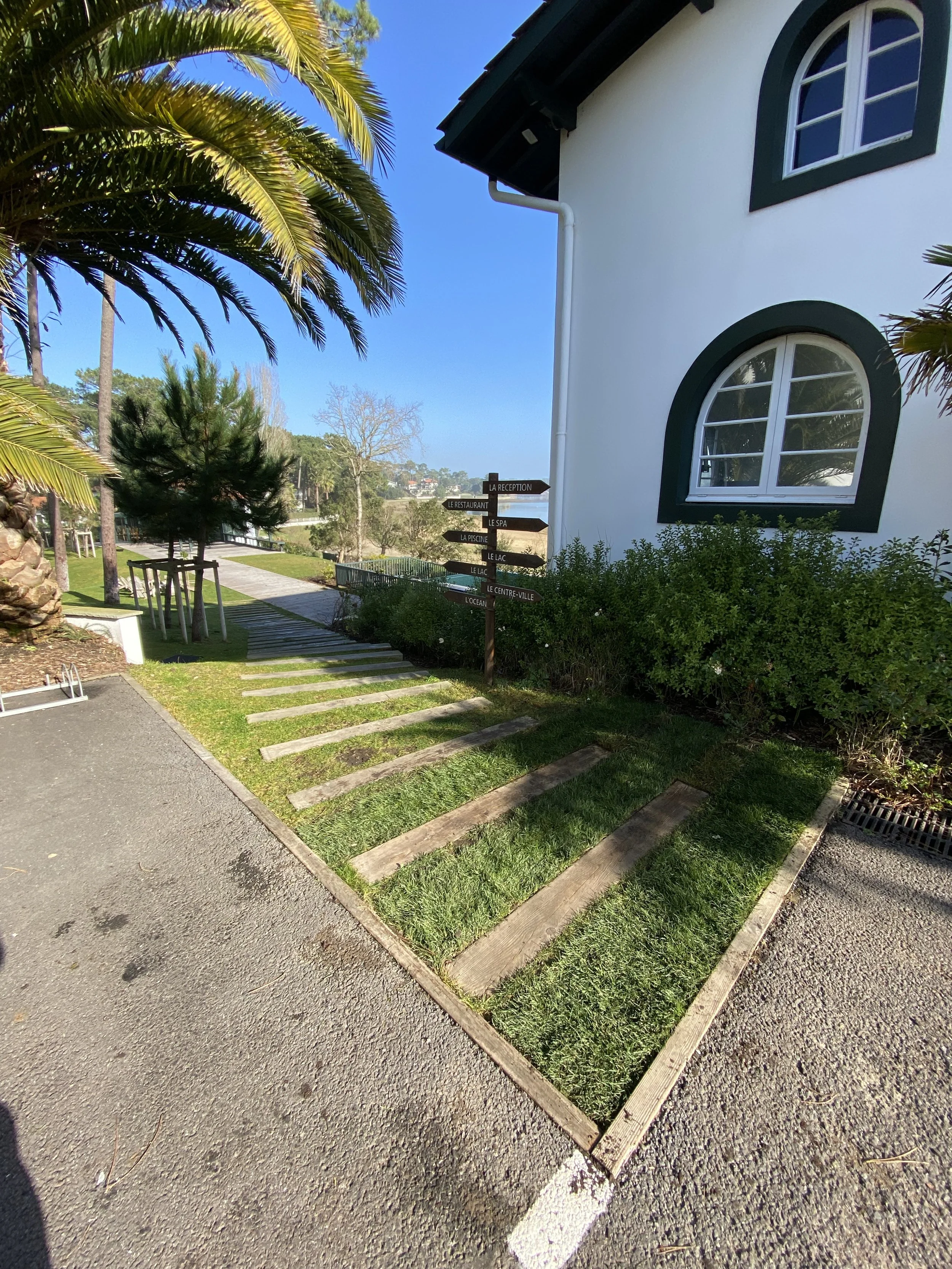 Une allée pavée bordée d'arbustes et d'arbres, devant une maison blanche avec des fenêtres à volets verts et un panneau indicateur en bois.