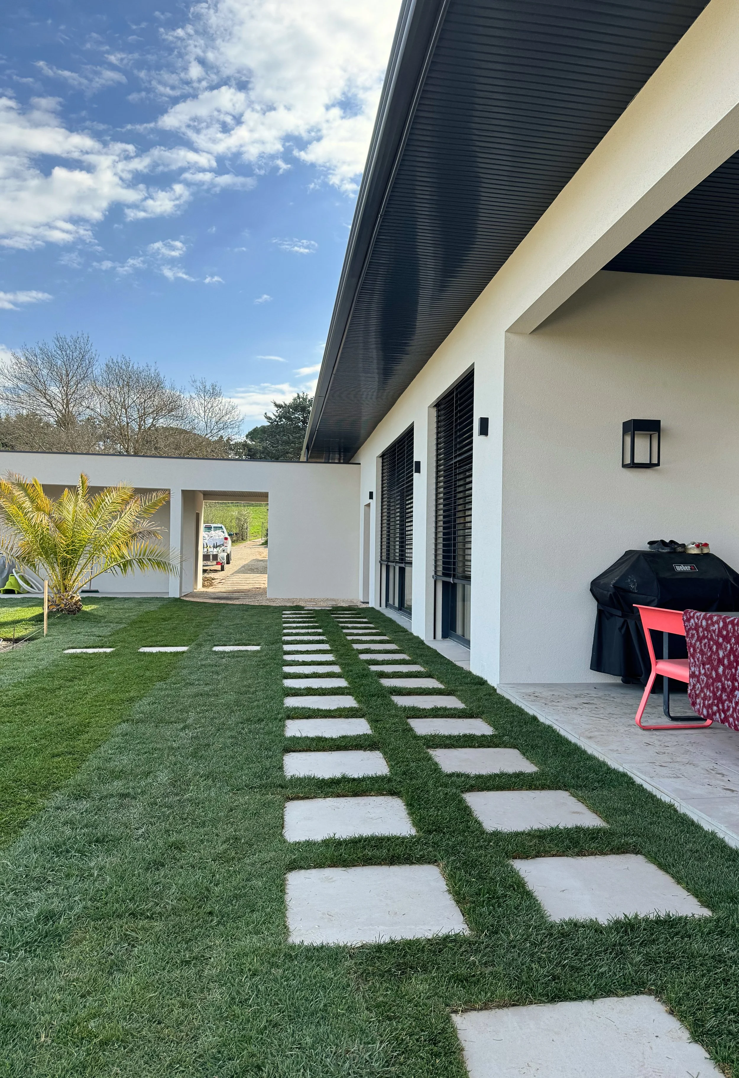 photo de la façade d'une maison moderne au style minimaliste, avec un chemin pavé traversant le jardin en gazon, un palmier sur la gauche, un barbecue recouvert, une table avec des chaises roses et un contre-jour en arrière-plan sous un ciel bleu avec quelques nuages.