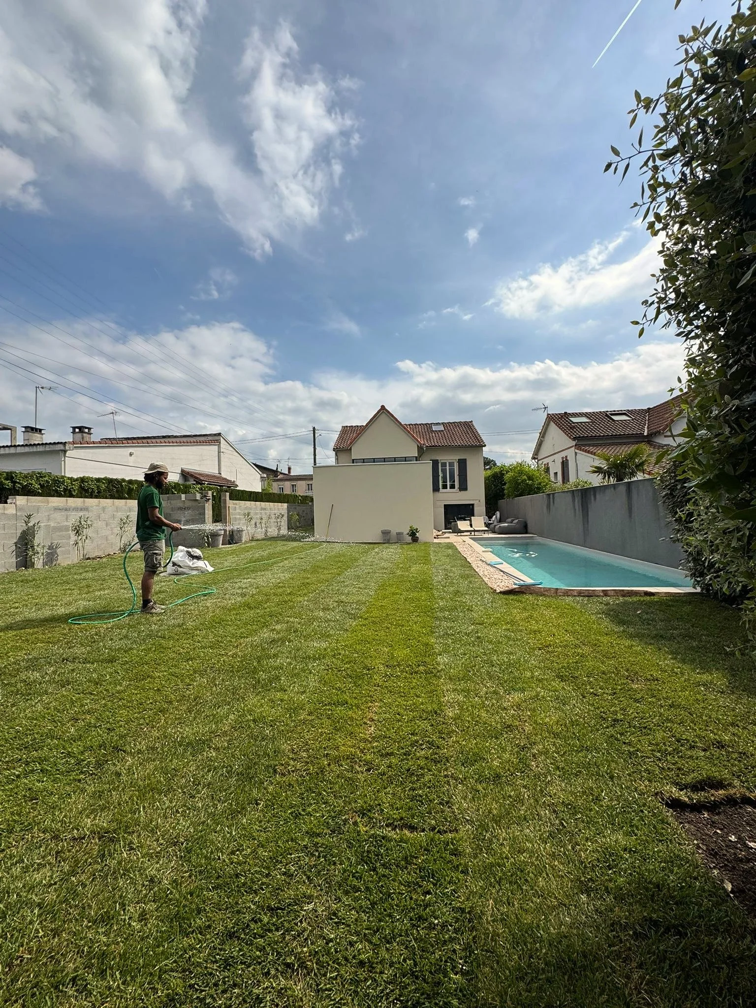 Une personne arrosant la pelouse dans un jardin résidentiel avec une piscine, des maisons en arrière-plan et un ciel partiellement nuageux.