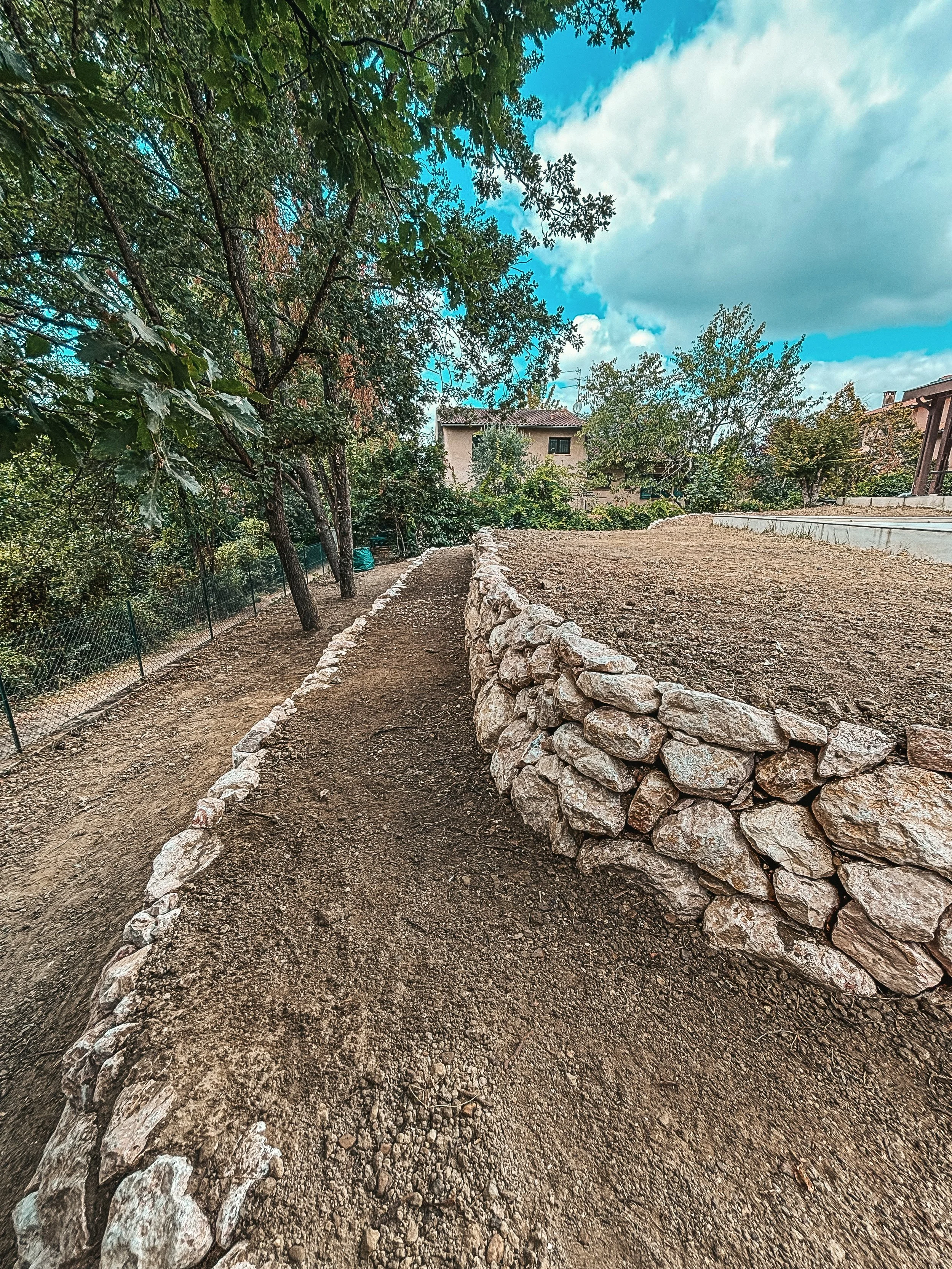 Une allée en terre bordée de pierres avec des arbres verts et un ciel nuageux en arrière-plan.
