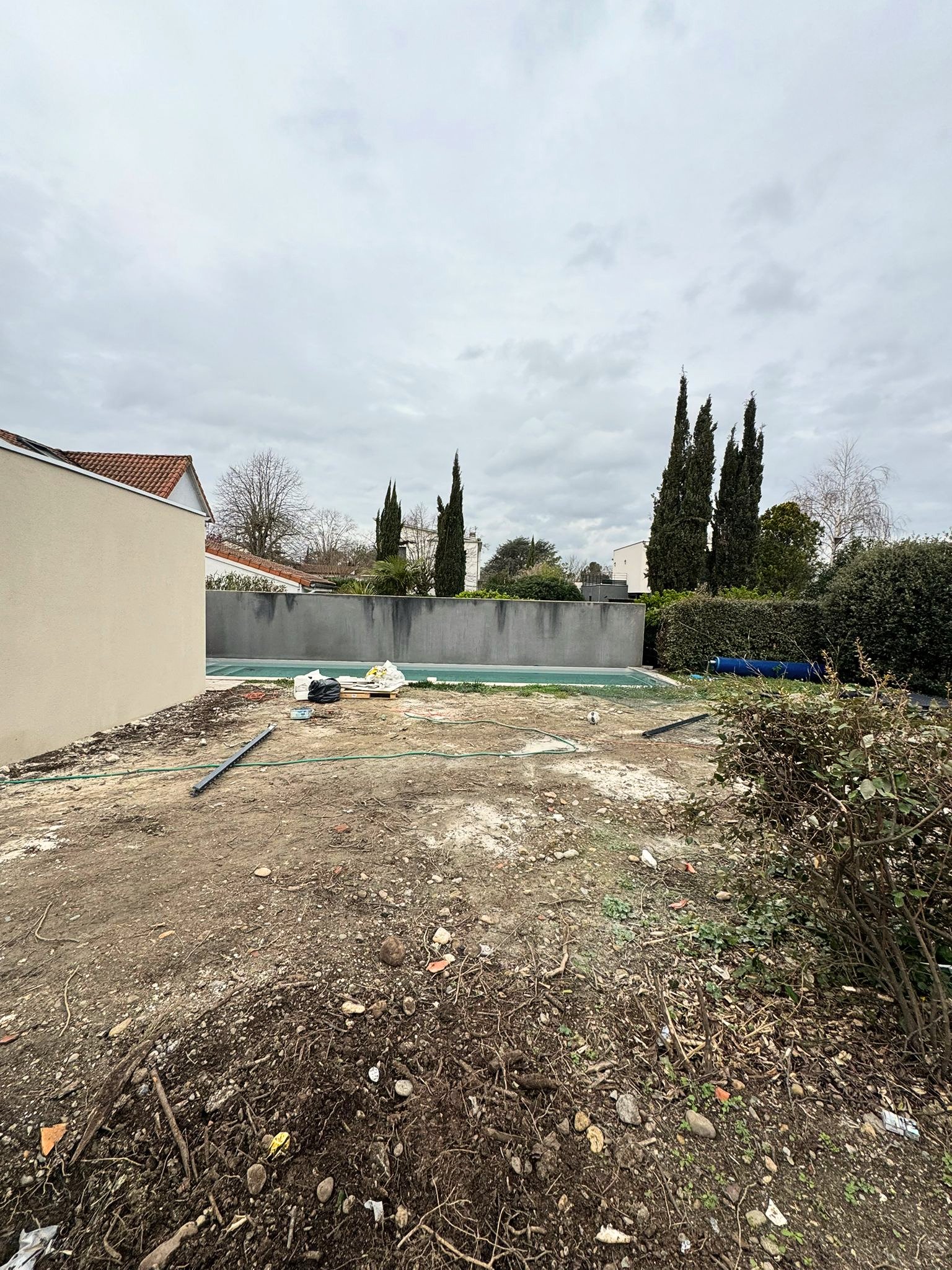 Un jardin en cours de rénovation avec sol déterré, pierres, branches et outils, entouré d'une clôture en béton et de jeunes arbres et arbustes.