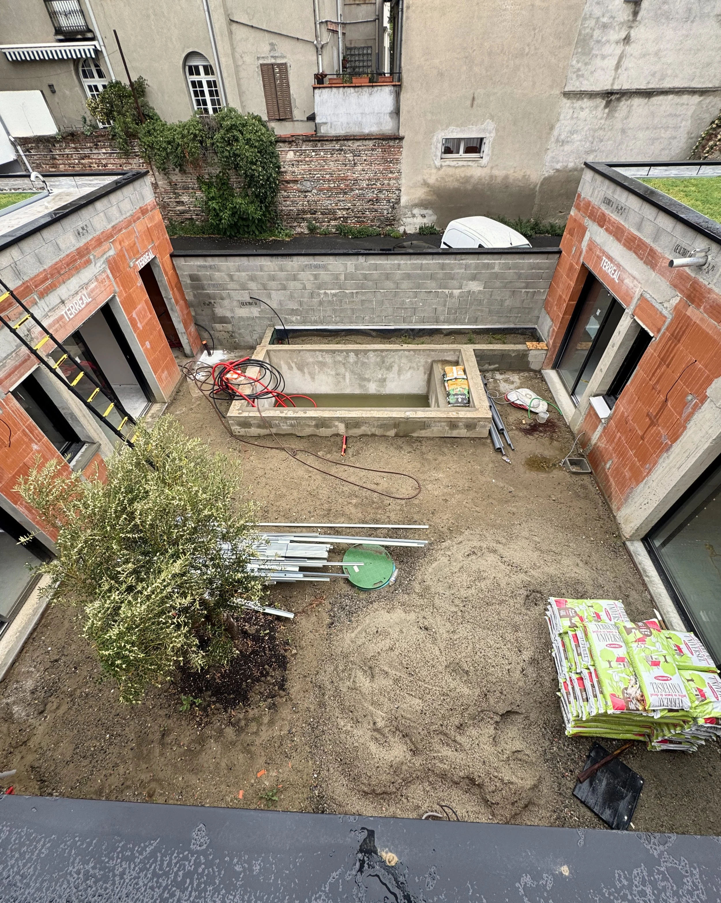 Vue d'une cour en cours d'aménagement avec un petit bassin en construction, des sacs de terreau empilés, une plante à gauche, et un bâtiment en construction sur la droite.