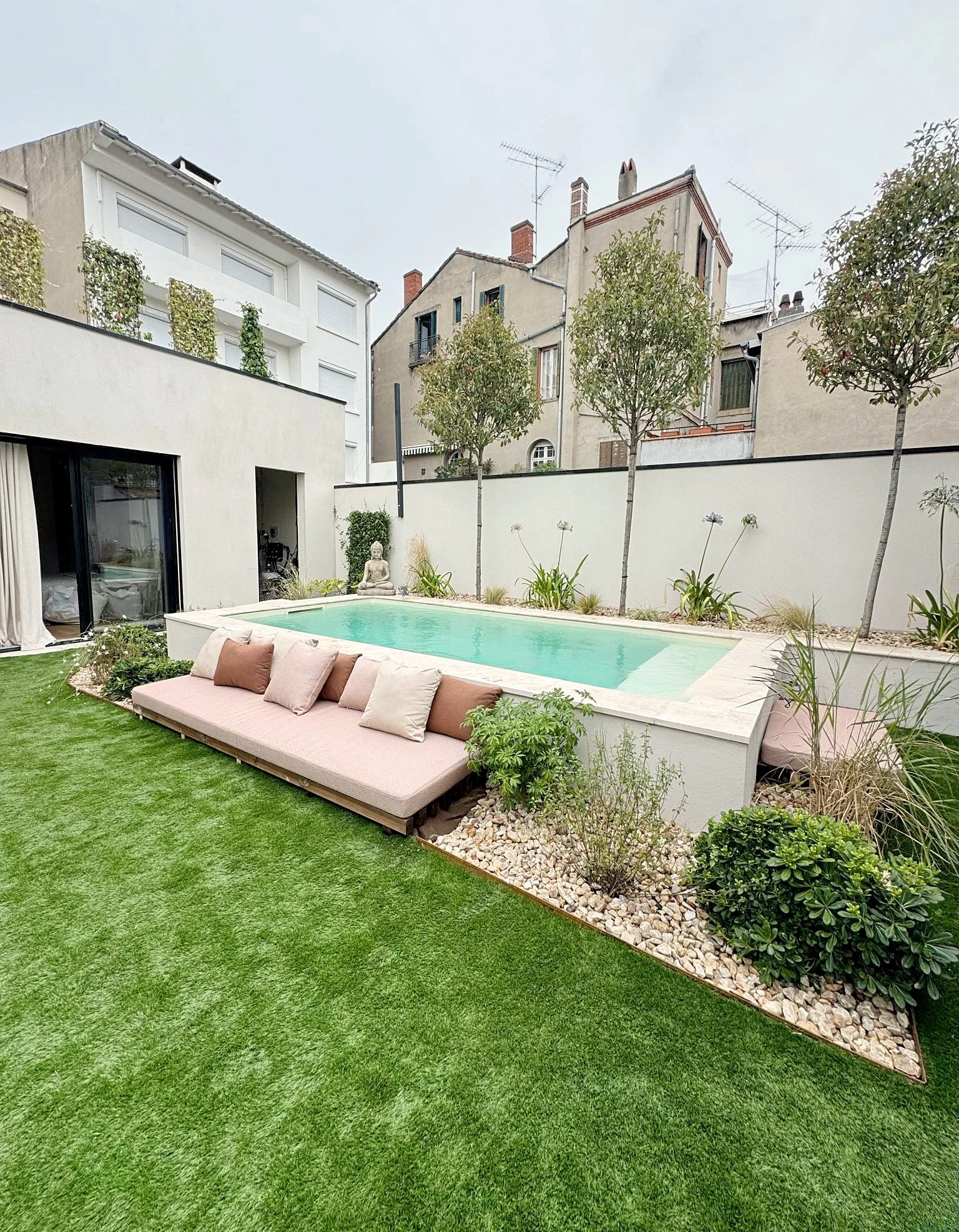 Un jardin avec une piscine, un canapé avec plusieurs coussins, et des plantes autour, avec un mur blanc en arrière-plan.