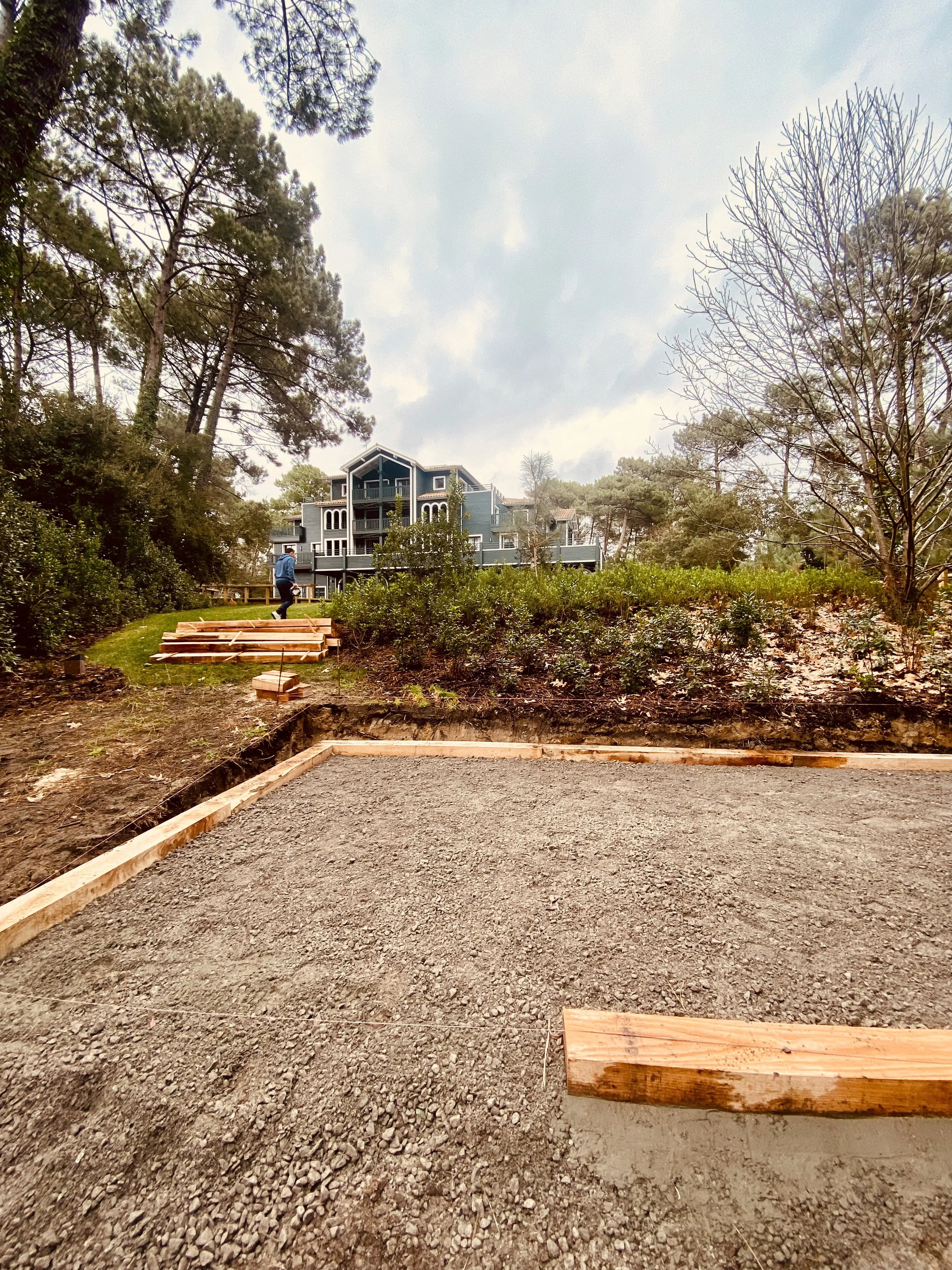 Une zone de construction avec le sol en gravier, des bords en bois, un arrière-plan avec une maison à plusieurs étages et des arbres entourant la zone.