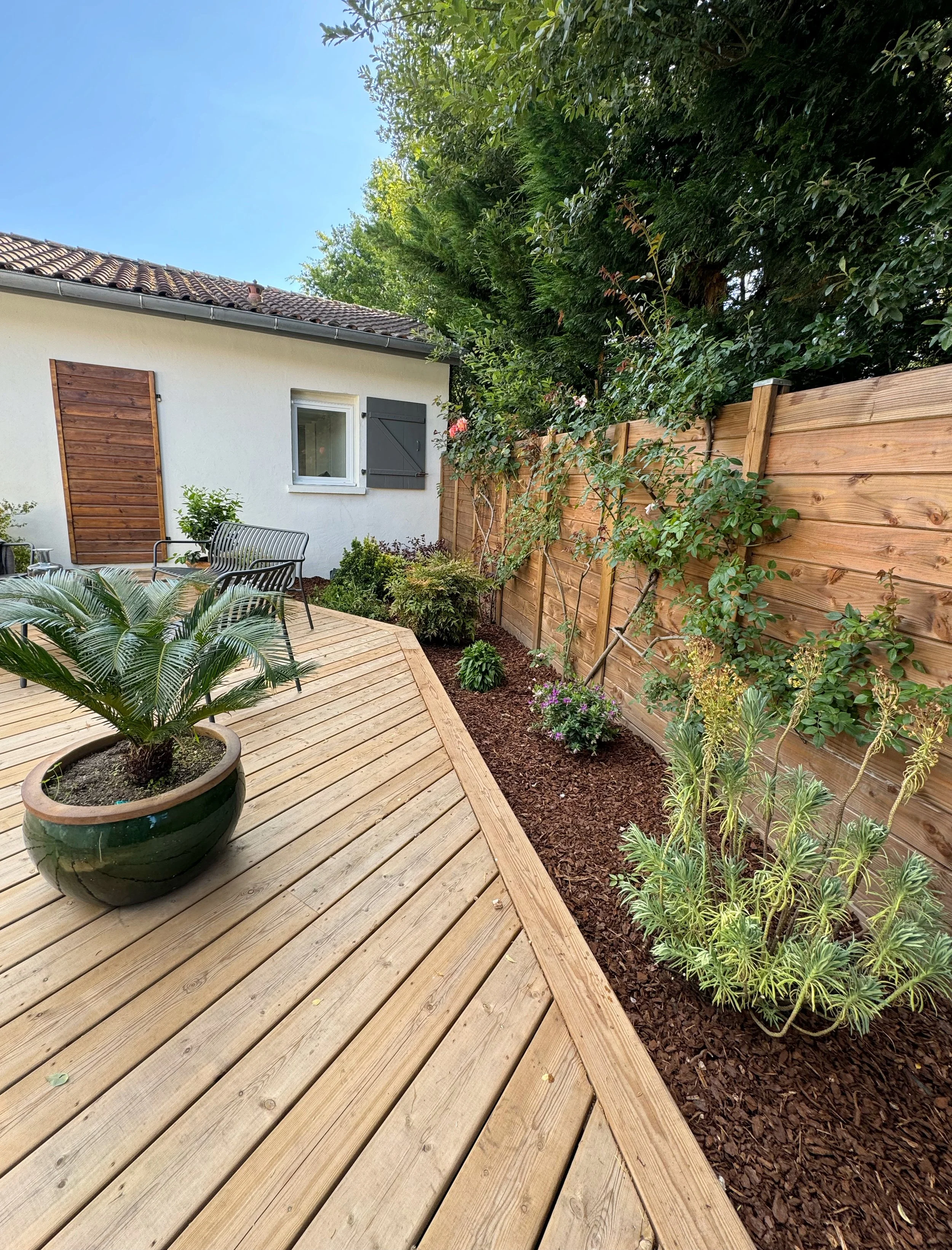Un jardin avec une terrasse en bois, des plantes en pot et une clôture en bois, avec un mur blanc et un toit en tuiles.