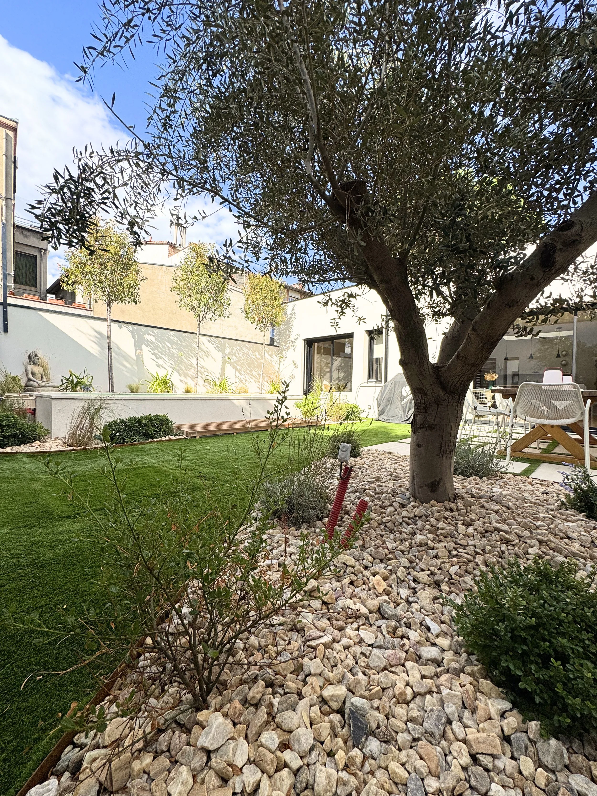 Un jardin avec un arbre au centre, entouré de pierre et d'herbe, avec un bâtiment moderne en arrière-plan et un ciel partiellement nuagé.