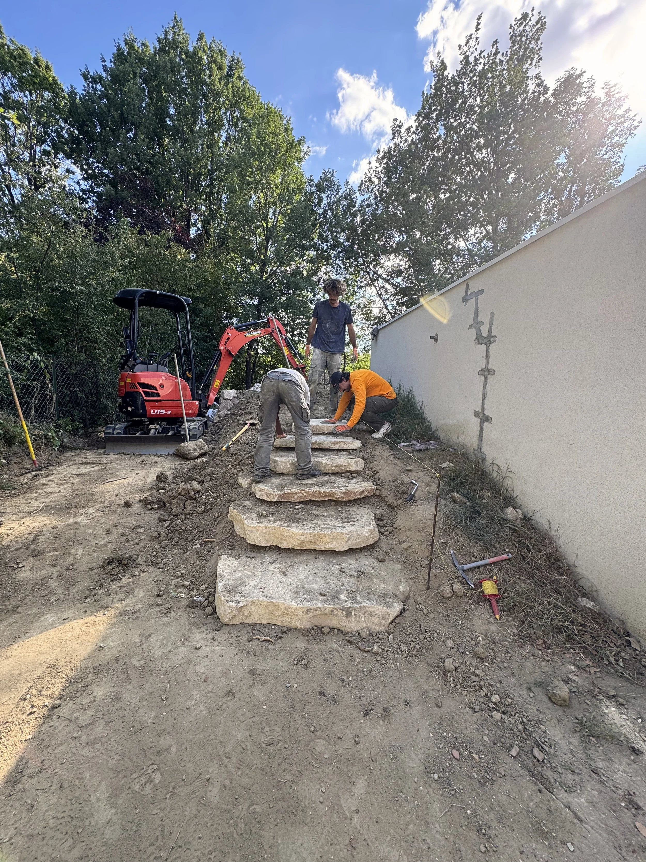 Trois hommes posent des escaliers en pierre dans un jardin, avec un petit excavateur rouge à côté, ensoleillé avec un ciel bleu et des arbres en arrière-plan.