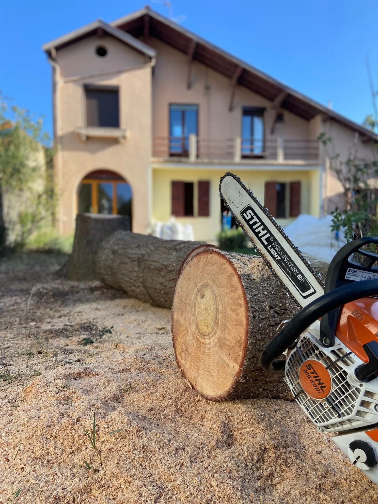 Une tronçonneuse à chaîne posée sur une partie coupée d'un arbre, avec une maison à deux étages en arrière-plan, un mur jaune, des fenêtres et un balcon en bois.