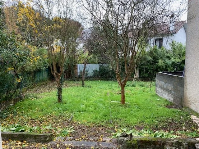 Un jardin verdoyant avec quelques arbres, entouré de clôtures et de maisons visibles en arrière-plan.
