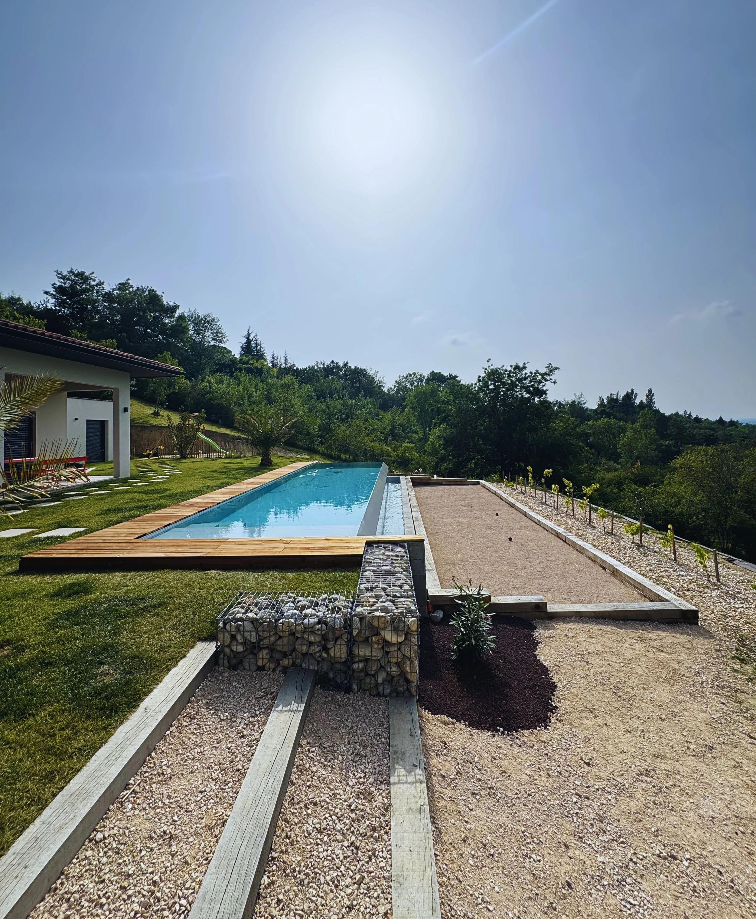 Piscine en plein air avec jardin, maison moderne, arbres et ciel ensoleillé.