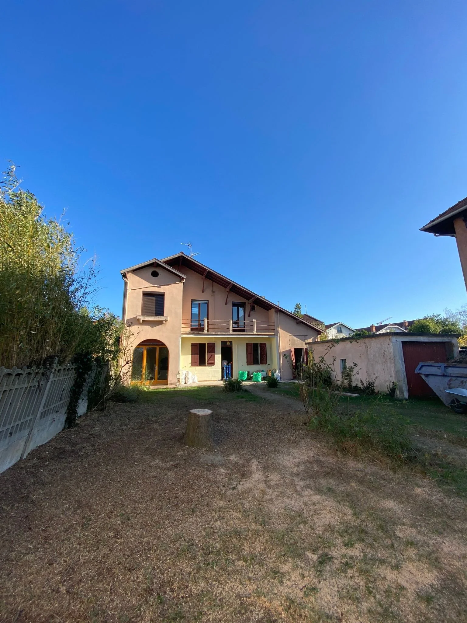 Vue d'une cour arrière avec une maison beige à deux étages, un arbre sur la gauche, un espace de jardin avec du sol en terre et peu de végétation, en fin de journée.